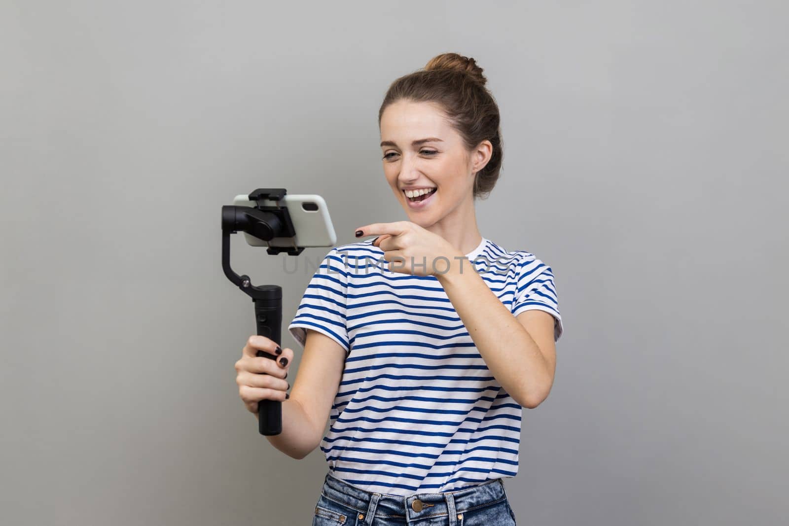 Portrait of attractive woman wearing striped T-shirt pointing to cell phone camera with smile, streaming, talking with followers, uses steadicam. Indoor studio shot isolated on gray background.