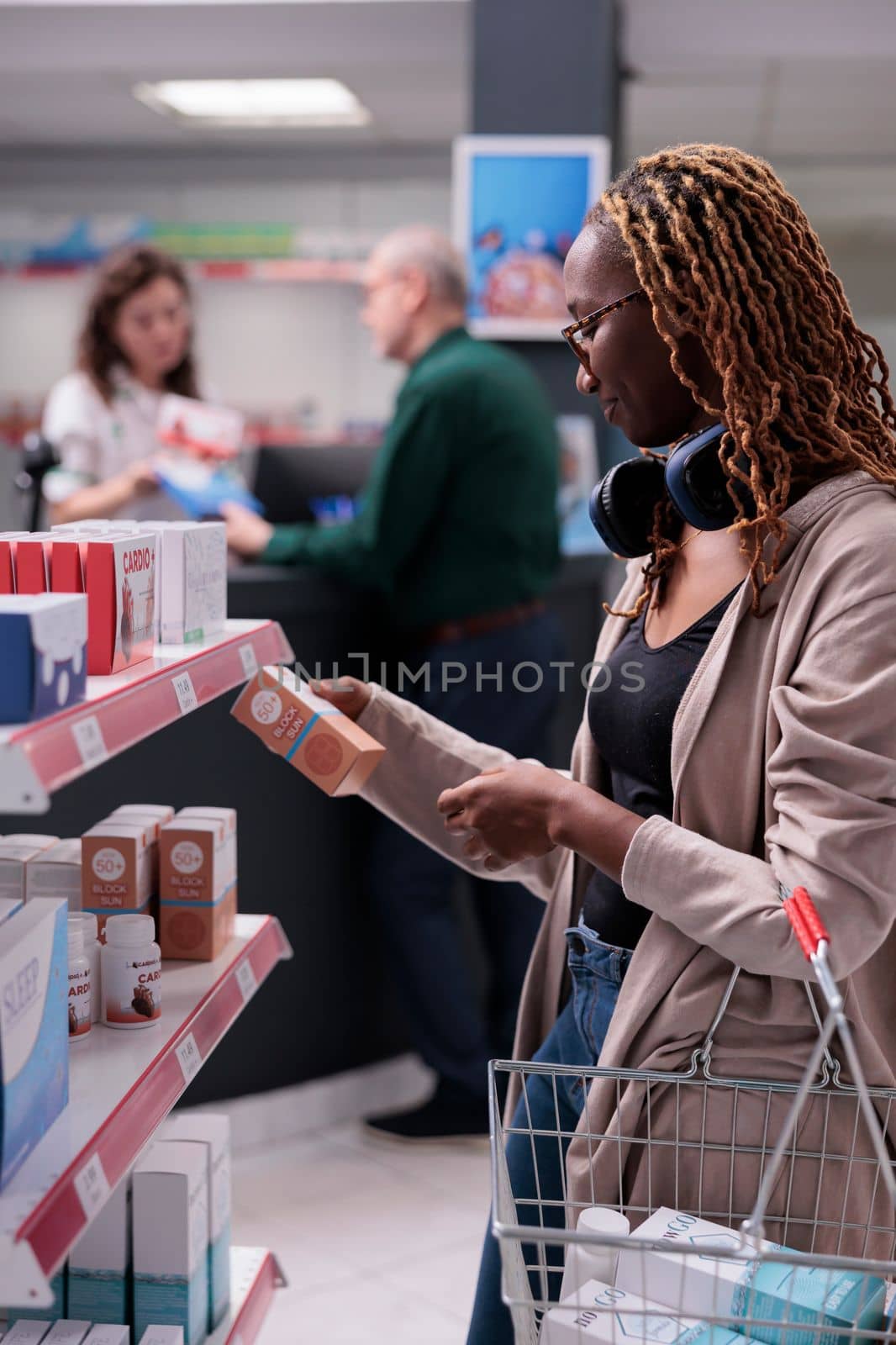 Customer looking at shelves full with pharmaceutical products, buying supplements after reading medication leaflet. Clients holding baskets putting pills package in it. Health care service