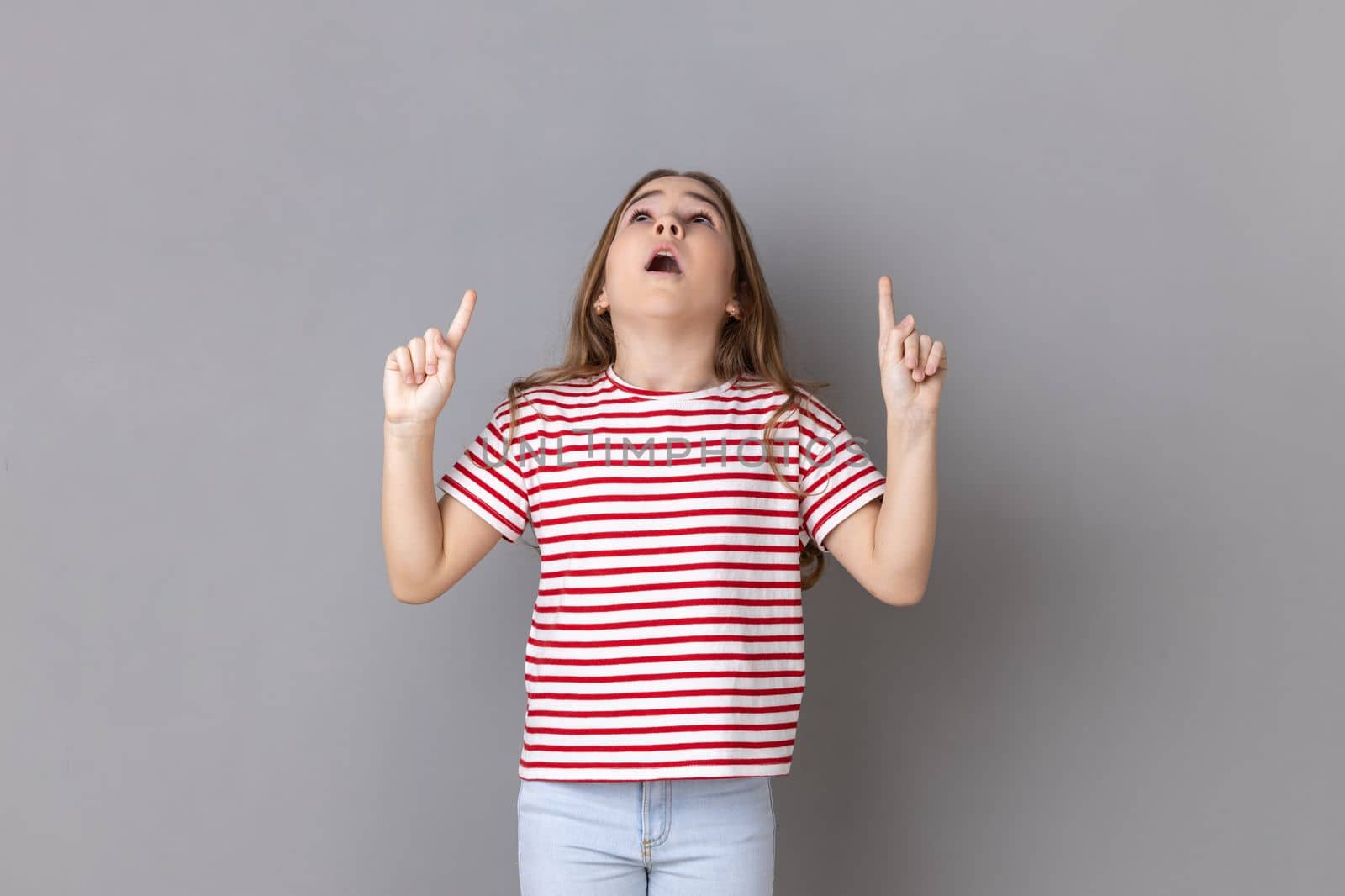 Wow look there, advertising area. Little girl looking astonished and pointing up copy space, showing empty place above head for promotion. Indoor studio shot isolated on gray background.