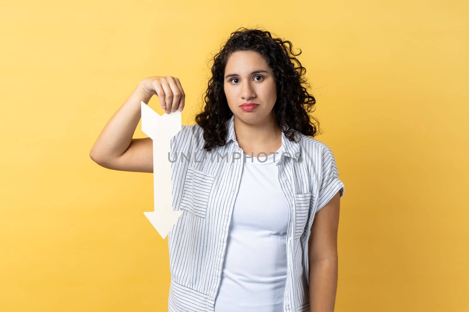 Portrait of upset dissatisfied woman with dark wavy hair showing white arrow pointing down, expressing sad emotions, downgrade concept. Indoor studio shot isolated on yellow background.