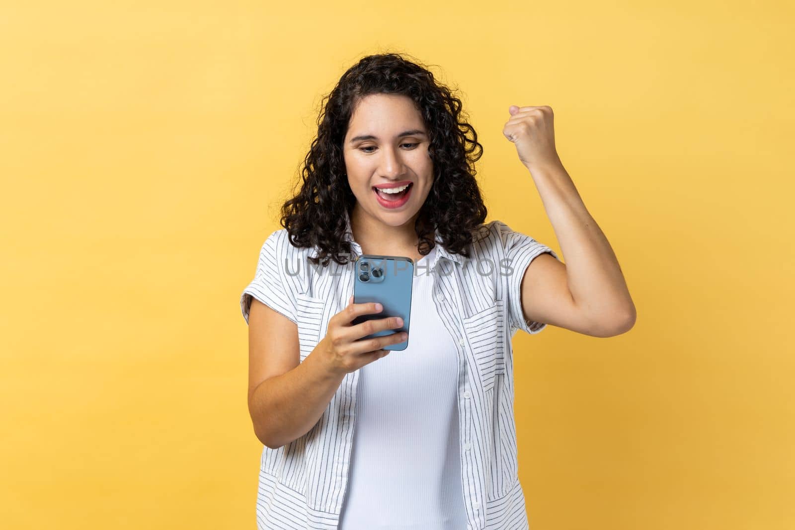 Portrait of extremely happy woman with dark wavy hair holding smart phone and clenched fist, showing yes gesture, celebrating her winning. Indoor studio shot isolated on yellow background.