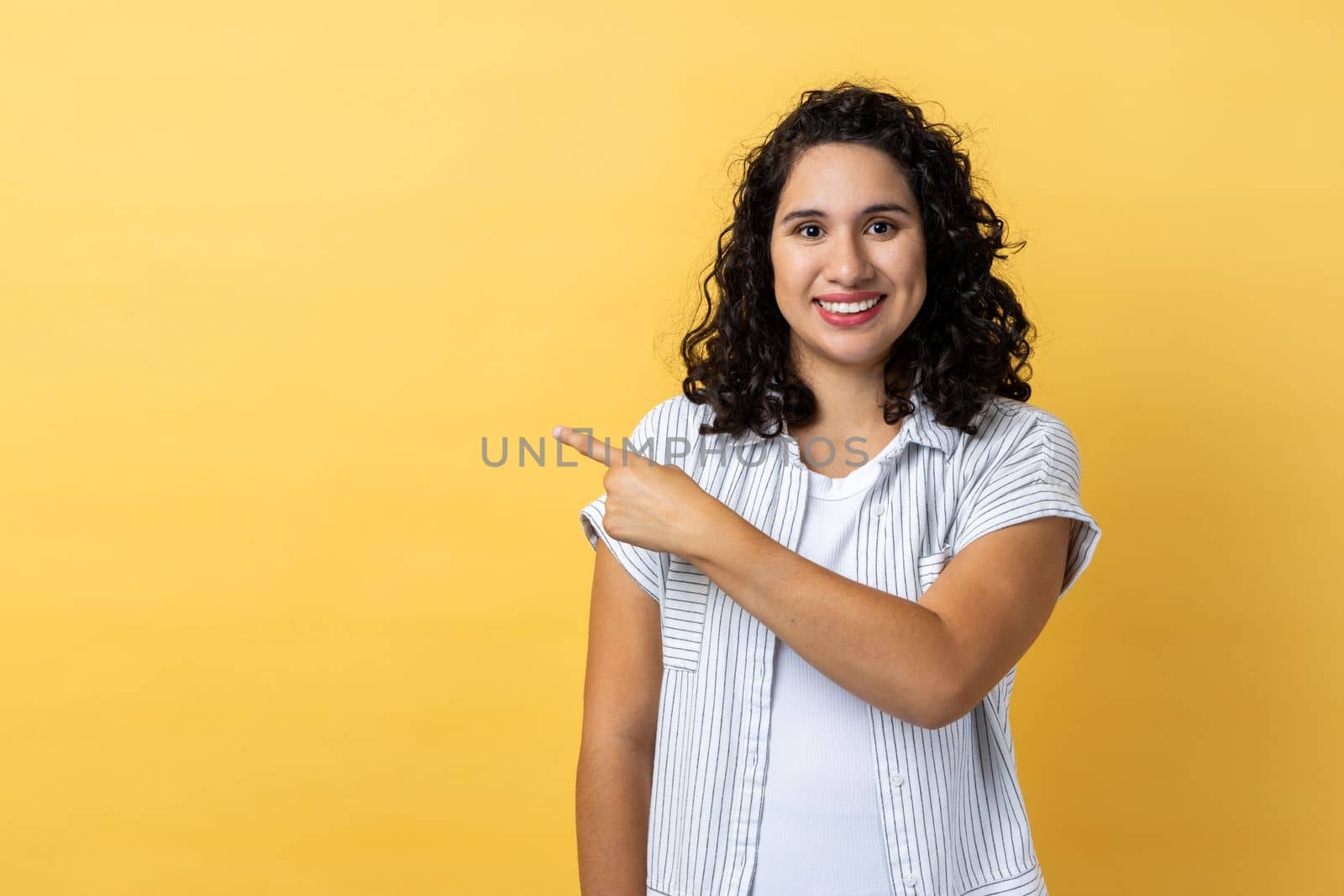 Portrait of smiling satisfied beautiful woman with dark wavy hair standing pointing aside with finger, showing copy space for advertisement. Indoor studio shot isolated on yellow background.