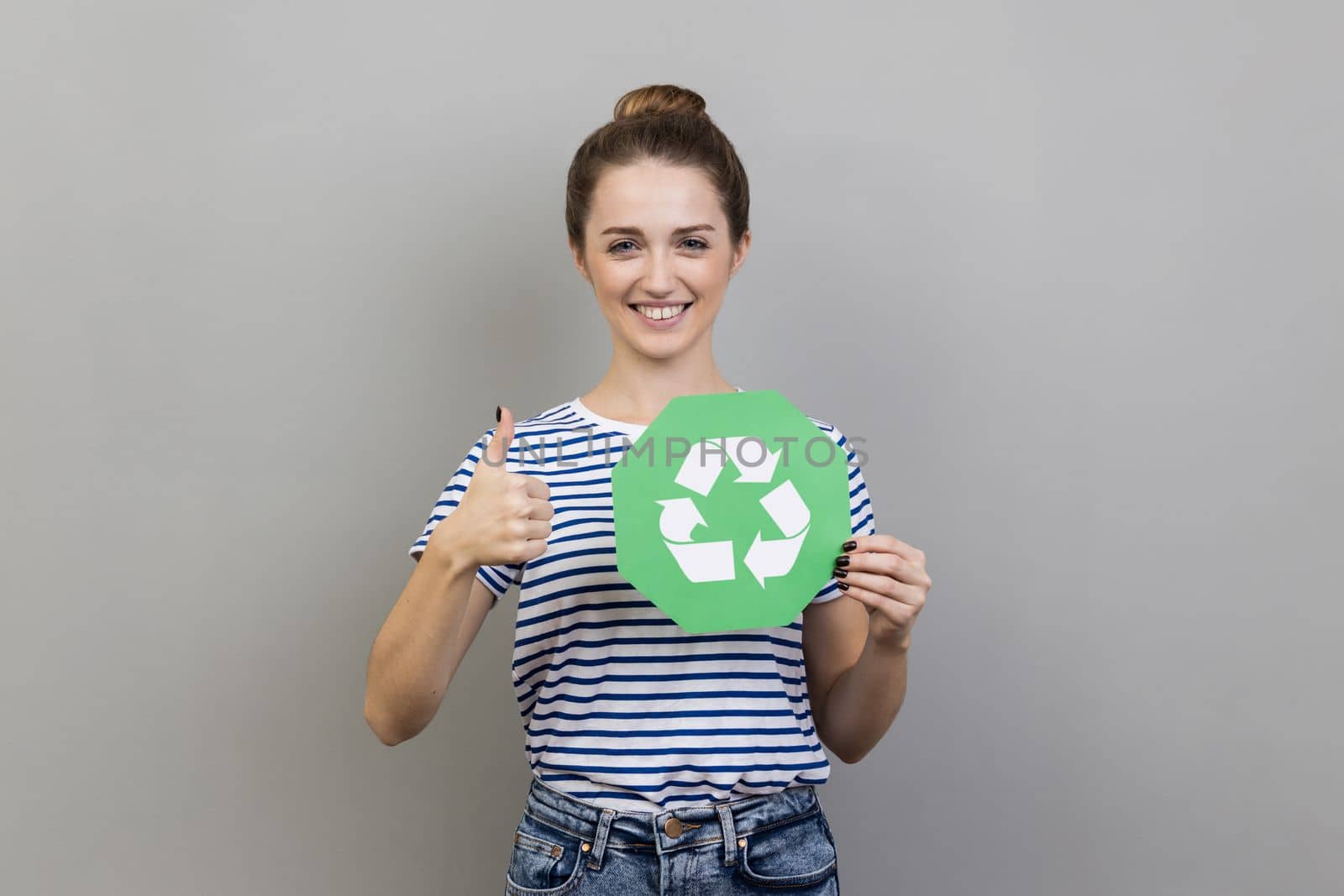 Portrait of smiling woman wearing striped T-shirt holding green recycling sign in hand and showing thumb up, like gesture, thinking green. Indoor studio shot isolated on gray background.