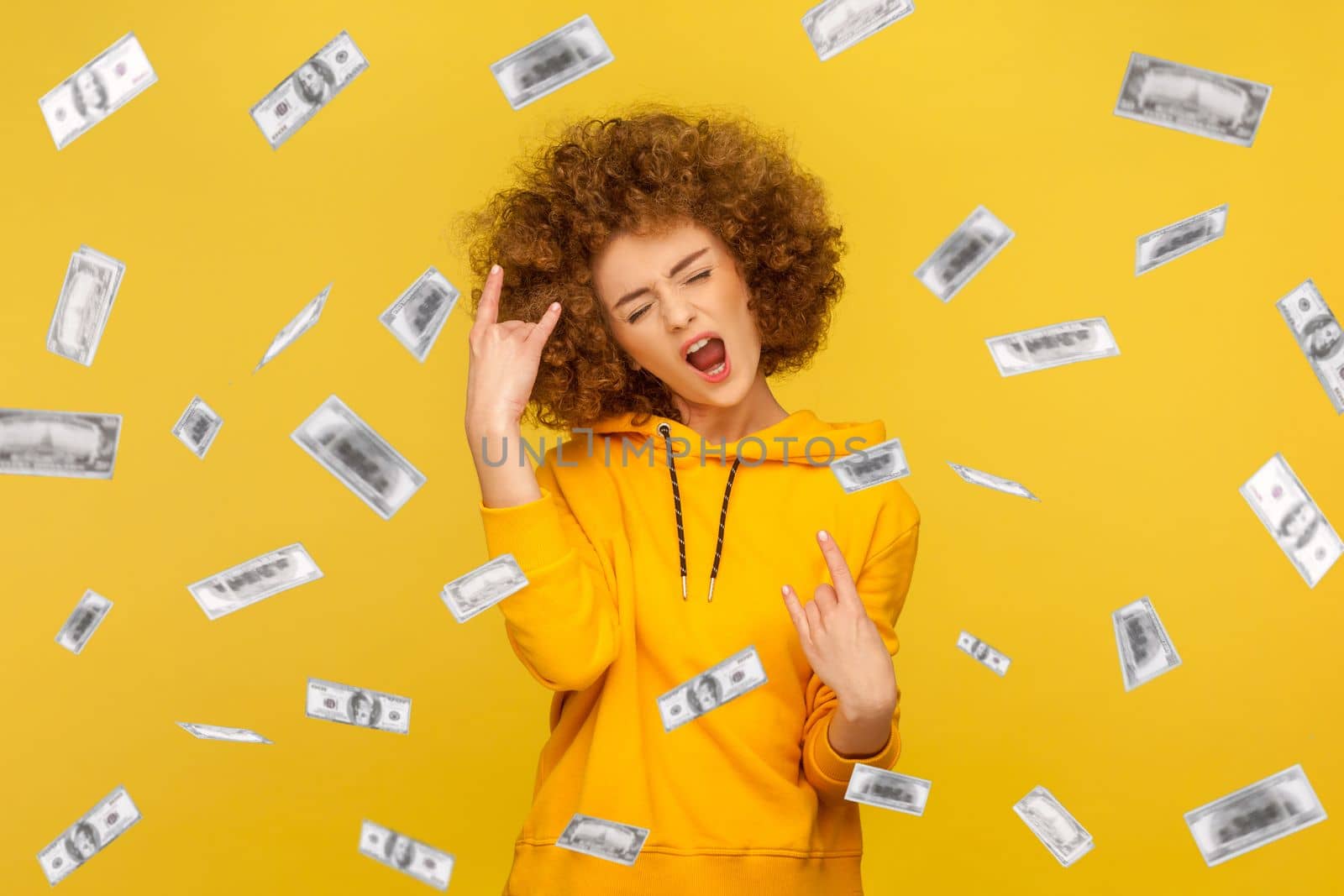 Portrait of woman with Afro hairstyle wearing casual style hoodie showing rock and roll, rejoicing money rain falling from up, winning lottery. Indoor studio shot isolated on yellow background.