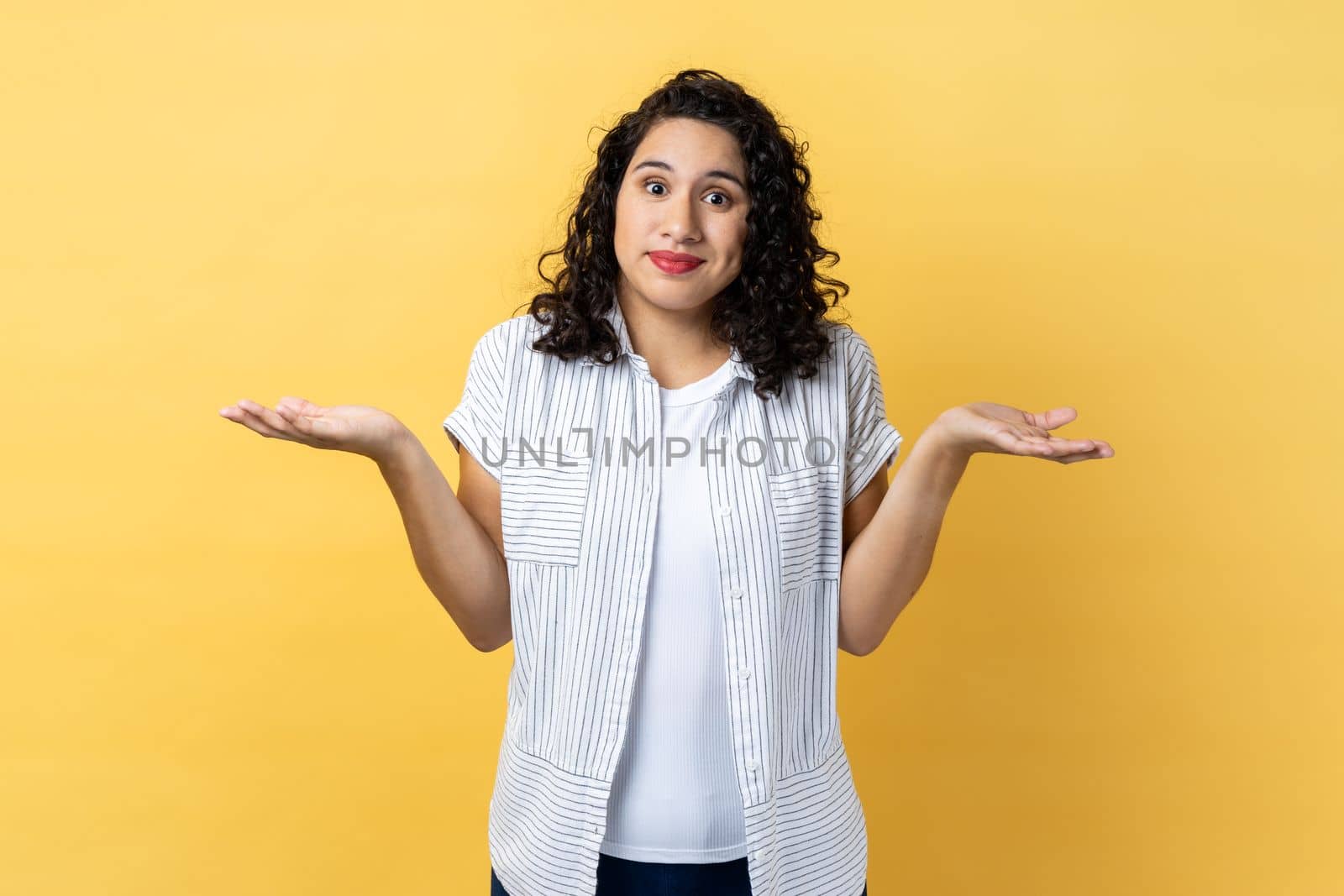I am not sure. Portrait of ambiguous woman with dark wavy hair standing with raised arms, looking away and don't know what to do. Indoor studio shot isolated on yellow background.