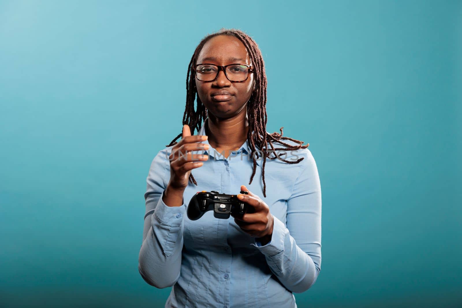 Sad young adult lady with modern gaming console controller standing on blue background. African american woman got upset because of lost online competitive videogame match.