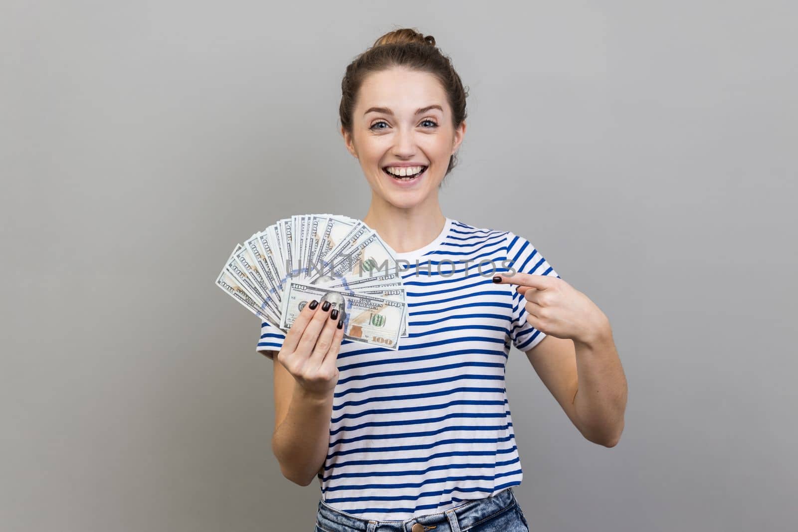 Portrait of amazed positive woman wearing striped T-shirt pointing at fan of dollar banknotes, showing big money and looking at camera. Indoor studio shot isolated on gray background.