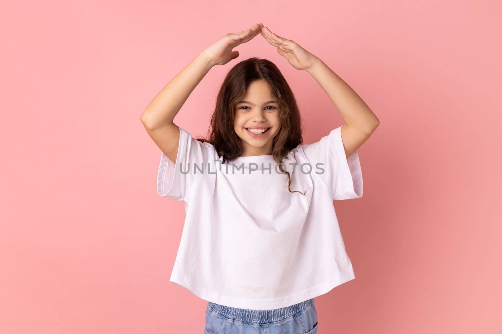 Portrait of little girl wearing white T-shirt with charming smile holding hands in gesture of house roof over head and smiling, feeling safe. Indoor studio shot isolated on pink background.