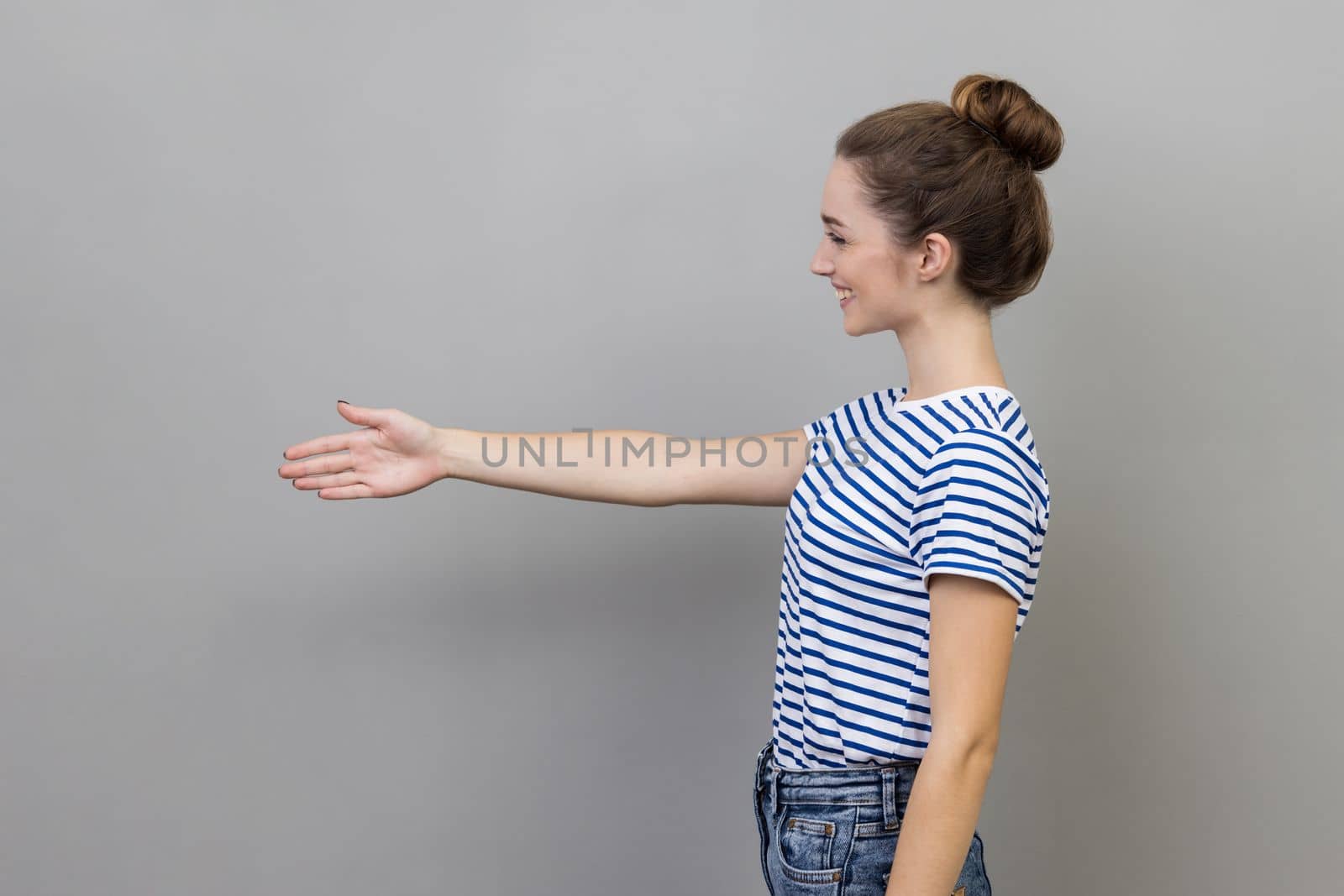 Side view of smiling friendly woman wearing striped T-shirt holding hand for handshake, happy to make agreement, successful partnership. Indoor studio shot isolated on gray background.