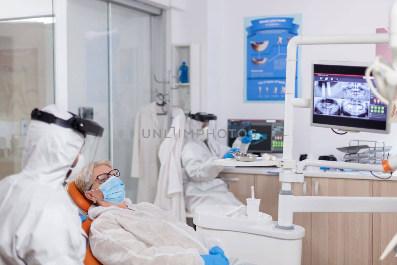 Dentist wearing protection agasint coronavirus during stomatology consultation of senior patient. Elderly woman in protective uniform during medical examination in dental clinic.