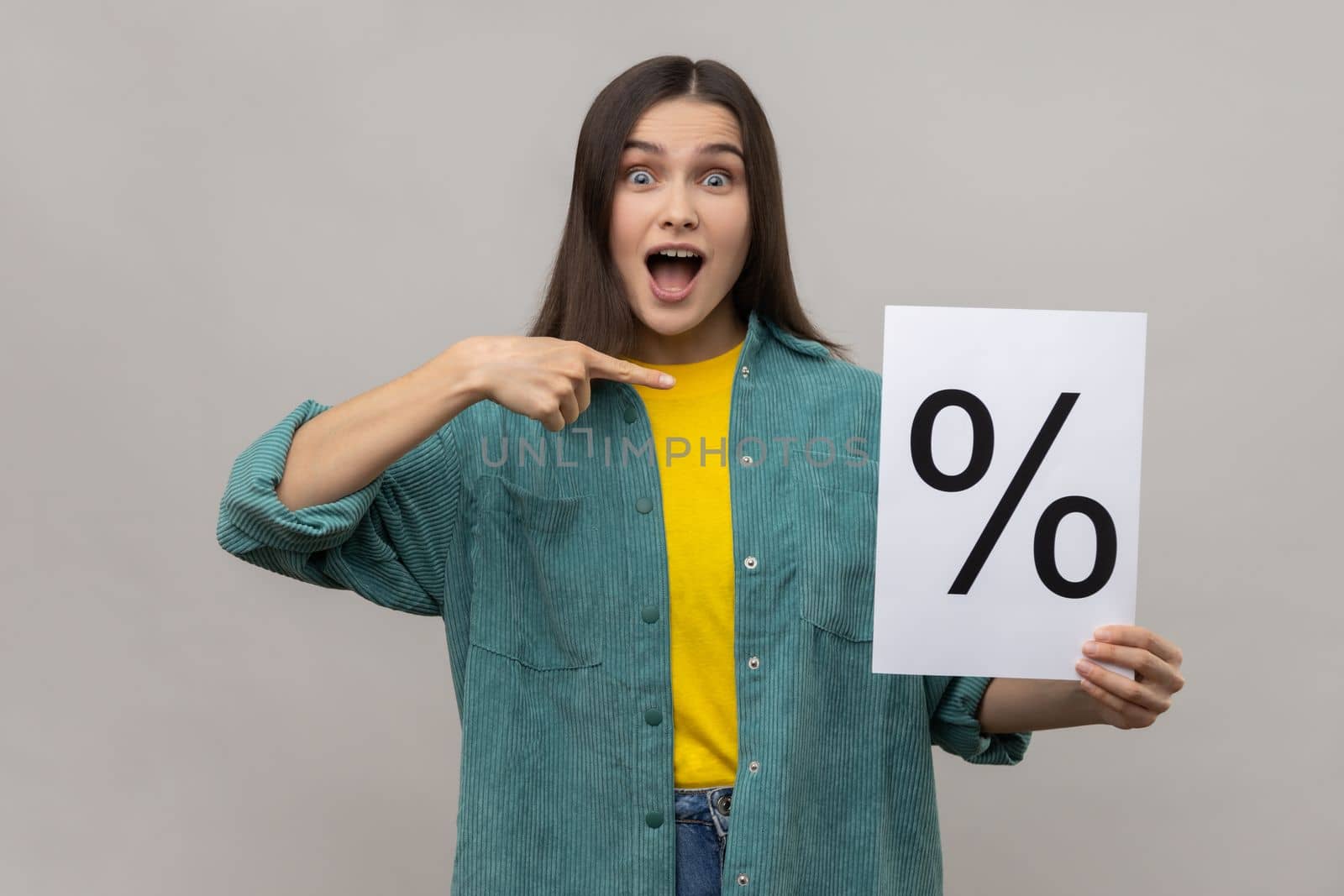 Amazed surprised woman holding and pointing at paper with percent sign inscription, looking at camera with open mouth, wearing casual style jacket. Indoor studio shot isolated on gray background.