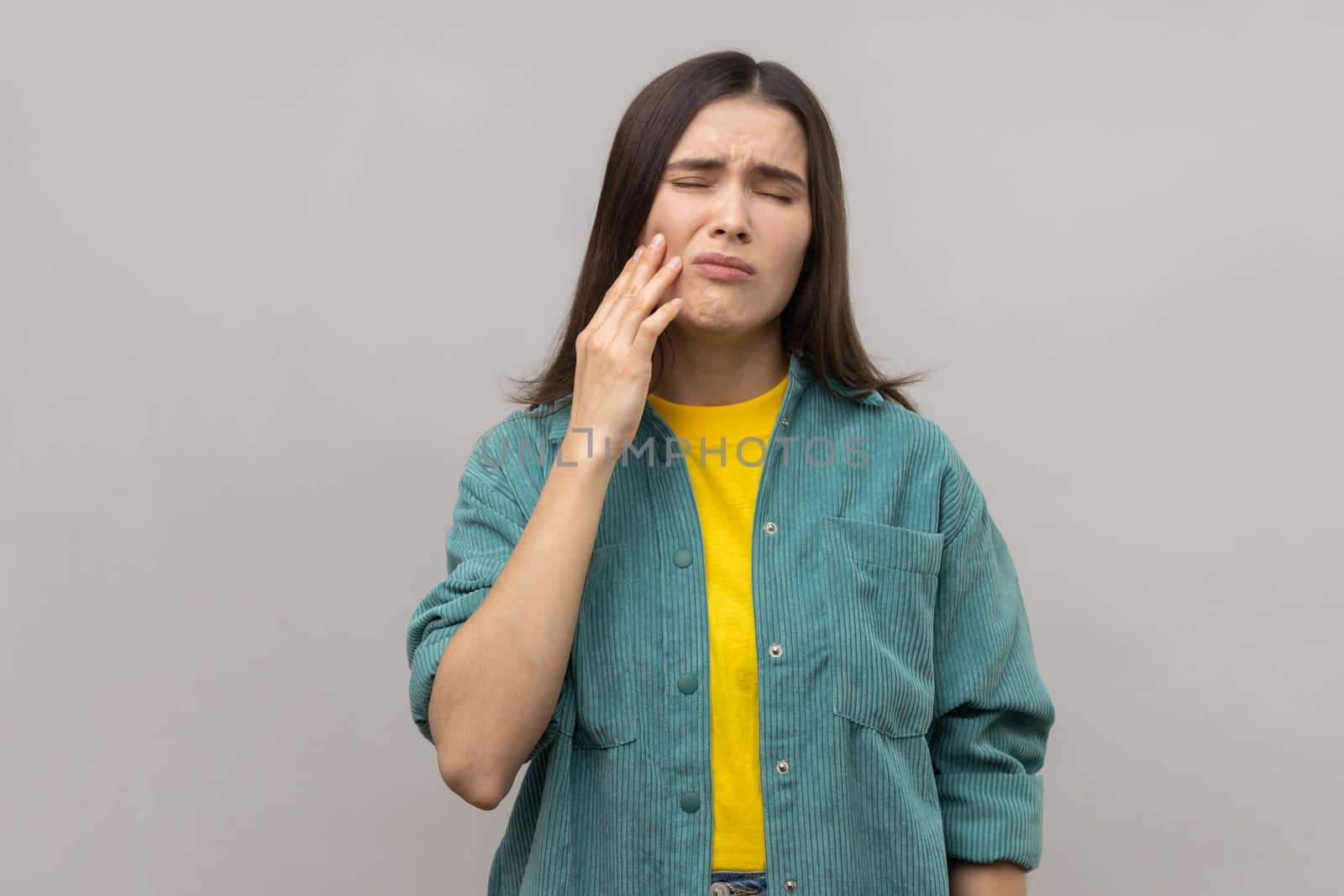 Unhealthy woman touching cheek, closing eyes with expression of terrible suffer from toothache, sensitive teeth, cavities, wearing casual style jacket. Indoor studio shot isolated on gray background.