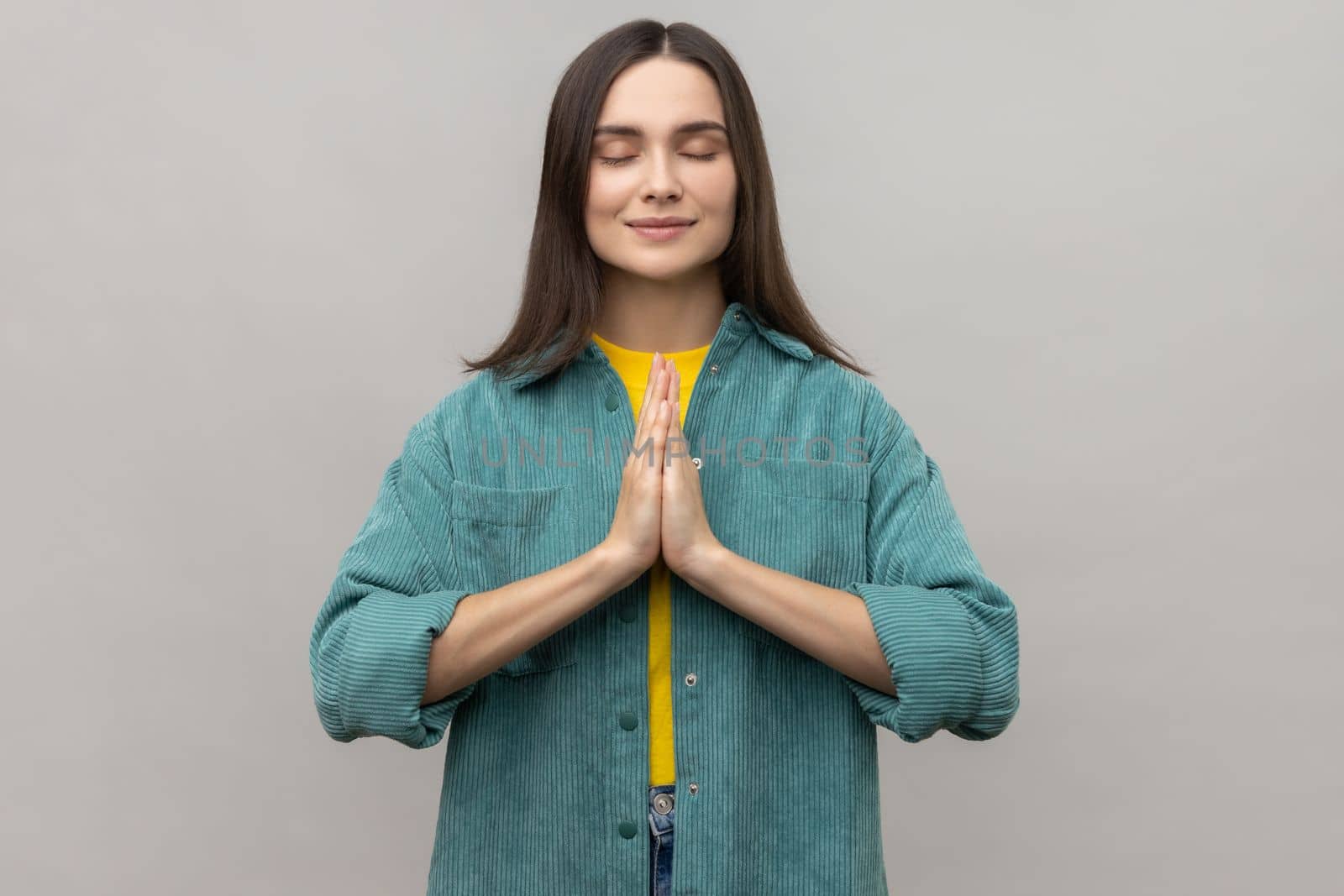 Gorgeous woman keeps hands in yoga gesture, has calm facial expression, keeping palms pressed together and closed eyes, wearing casual style jacket. Indoor studio shot isolated on gray background.