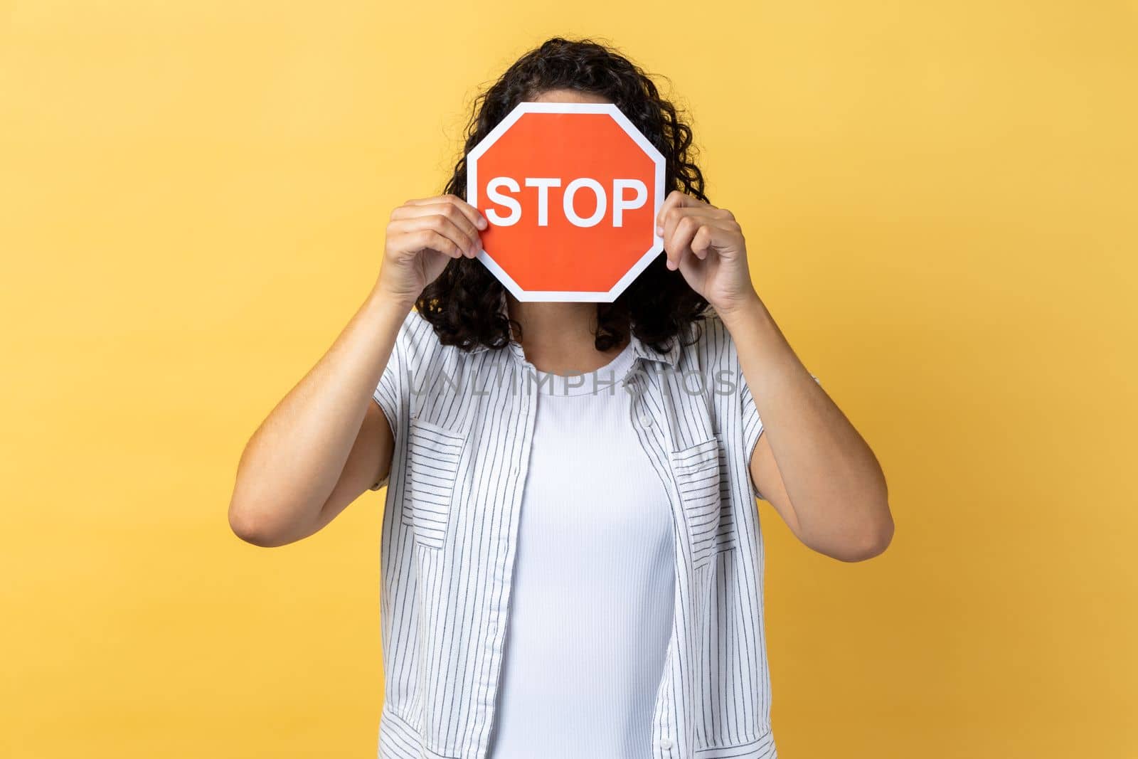 Anonymous woman with dark wavy hair standing covering her face with red stop sign, warning about problems, concept of ban, denial, prohibited to go. Indoor studio shot isolated on yellow background.