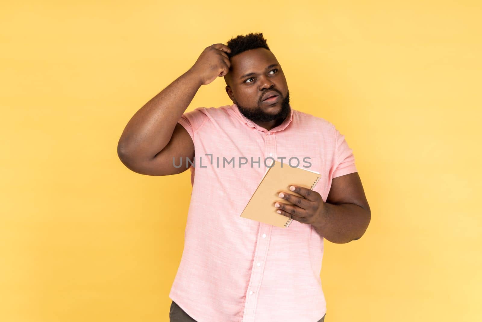 Portrait of thoughtful man wearing pink shirt thinking over smart idea, holding pencil and notebook to write plans, to-do list. Indoor studio shot isolated on yellow background.