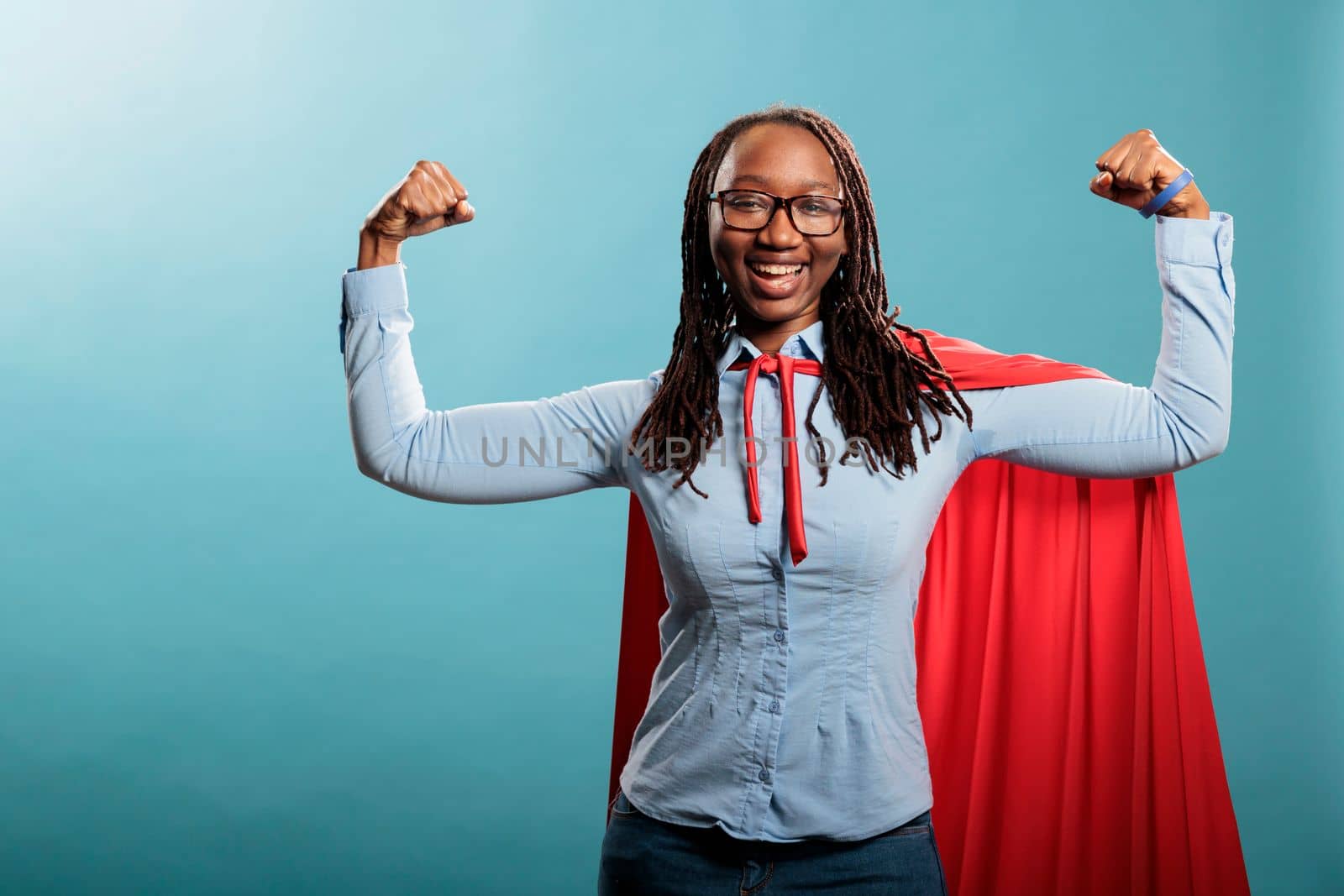 Happy positive justice defender flexing arms as a sign of strength while standing on blue background. Proud and tough looking young adult superhero woman showing empowerement and braveness.