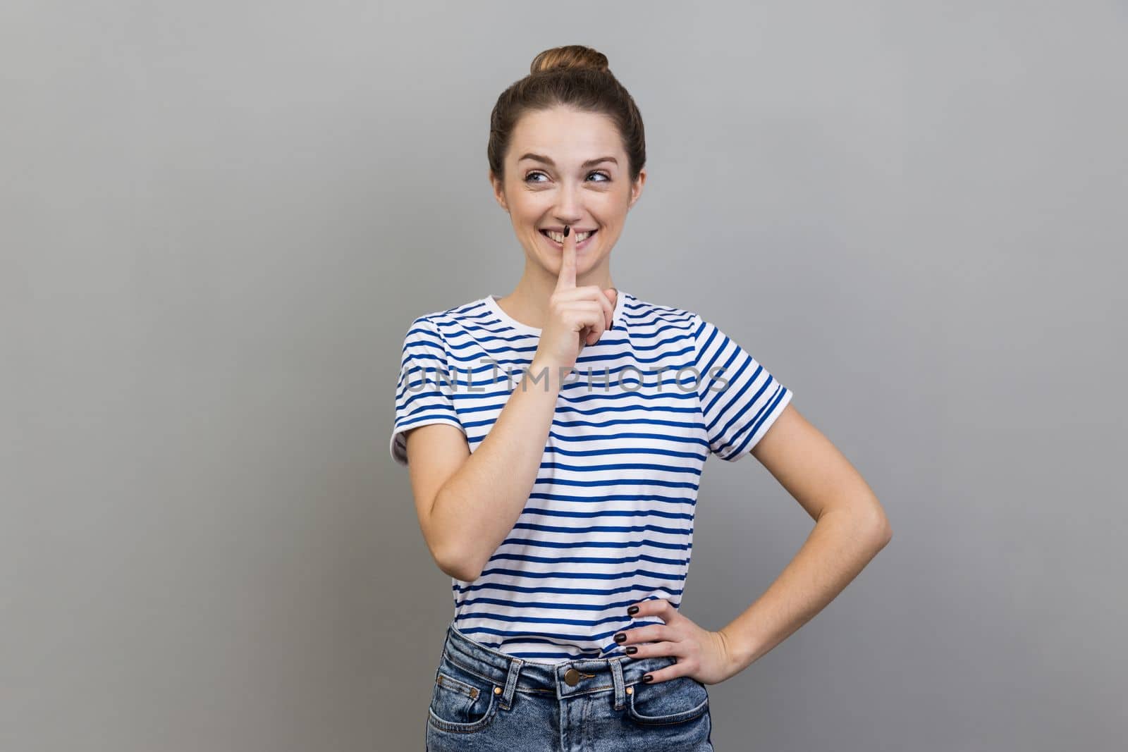 Portrait of woman wearing striped T-shirt showing shh gesture holding finger near lips, with naughty smile on face, making surprise, secret. Indoor studio shot isolated on gray background.