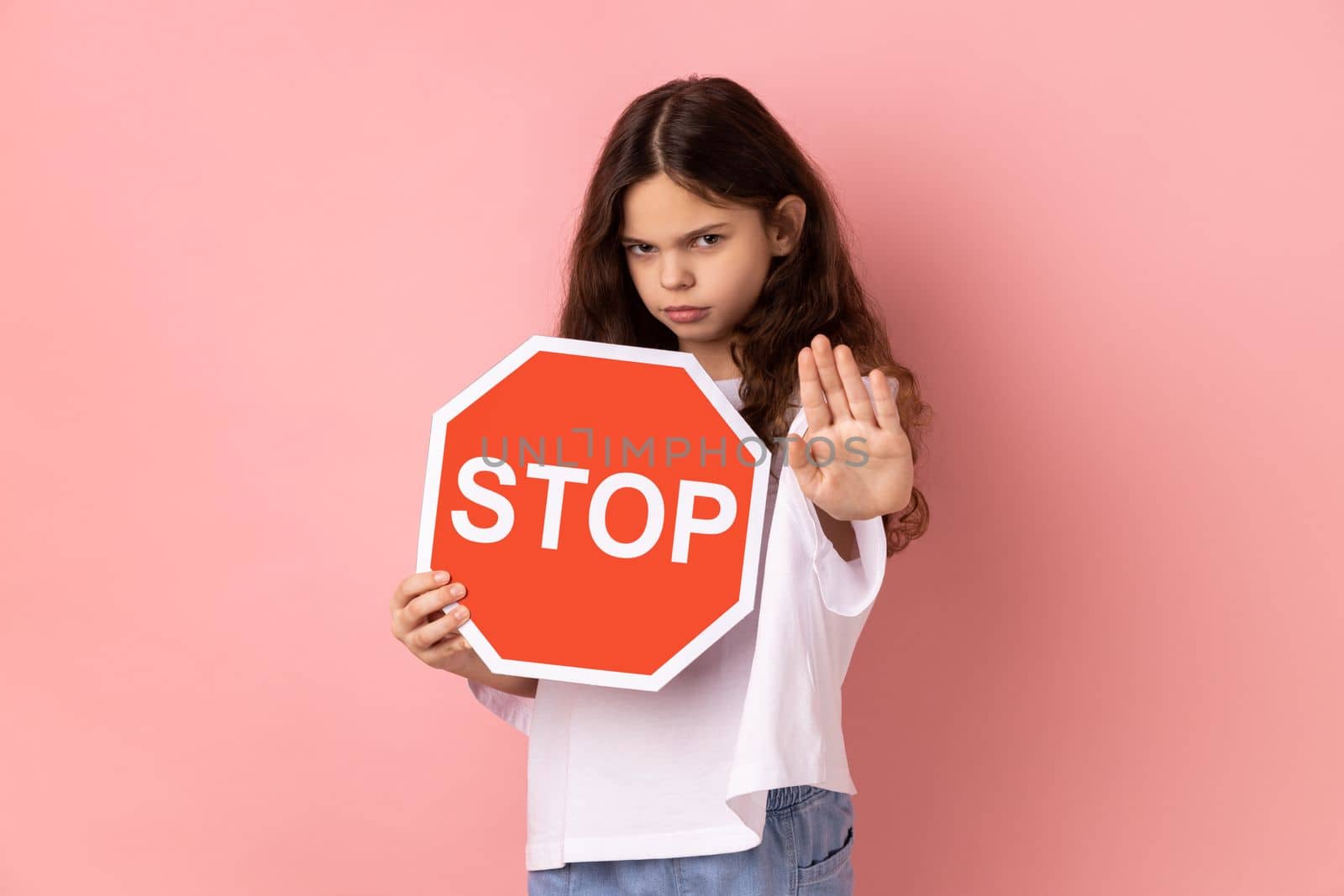 No, prohibited. Strict bossy little girl in T-shirt looking seriously at camera and showing stop gesture, holding road traffic sign as warning of ban. Indoor studio shot isolated on pink background.