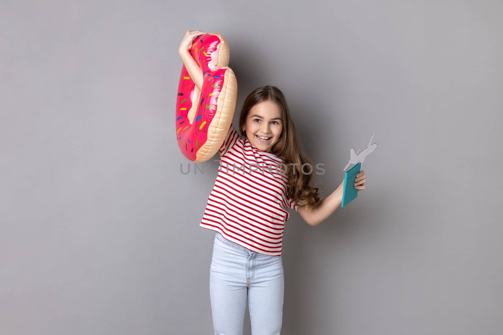 Portrait of excited satisfied little girl wearing striped T-shirt holding rubber ring and passport, being happy by future journey. Indoor studio shot isolated on gray background.