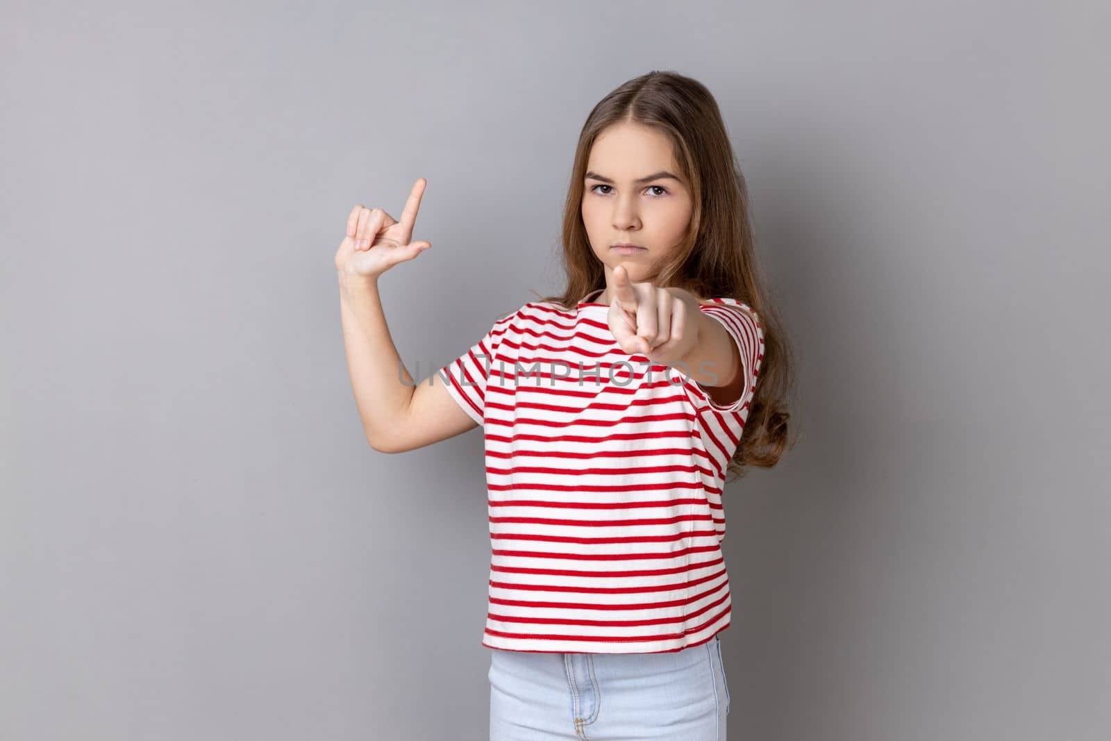 Portrait of serious little girl wearing striped T-shirt showing looser gesture and pointing at camera, looking with grumpy face. Indoor studio shot isolated on gray background.