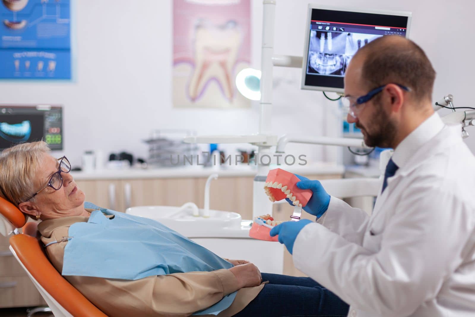 Dentist holding model of oral cavity with teeth during examination of senior patient. Medical teeth care taker discussing with senior woman about mouth hygiene.