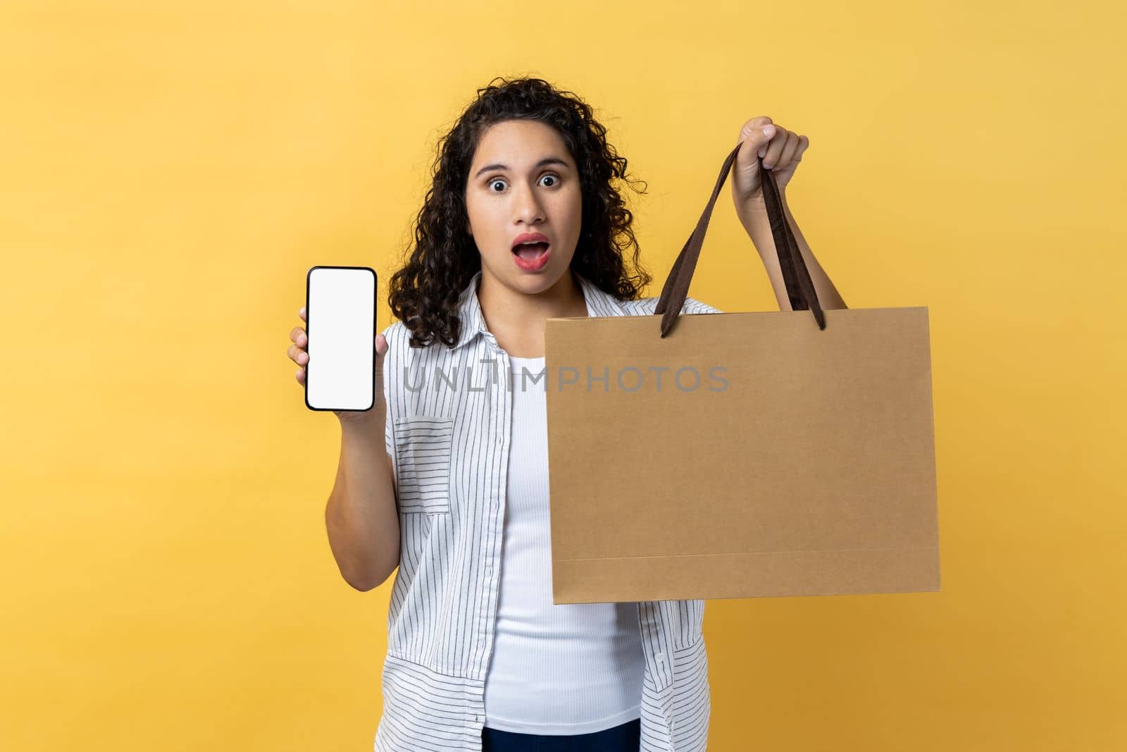 Portrait of surprised astonished woman with dark wavy hair holding paper shopping bag and mobile phone with empty screen, copy space for promotion. Indoor studio shot isolated on yellow background.