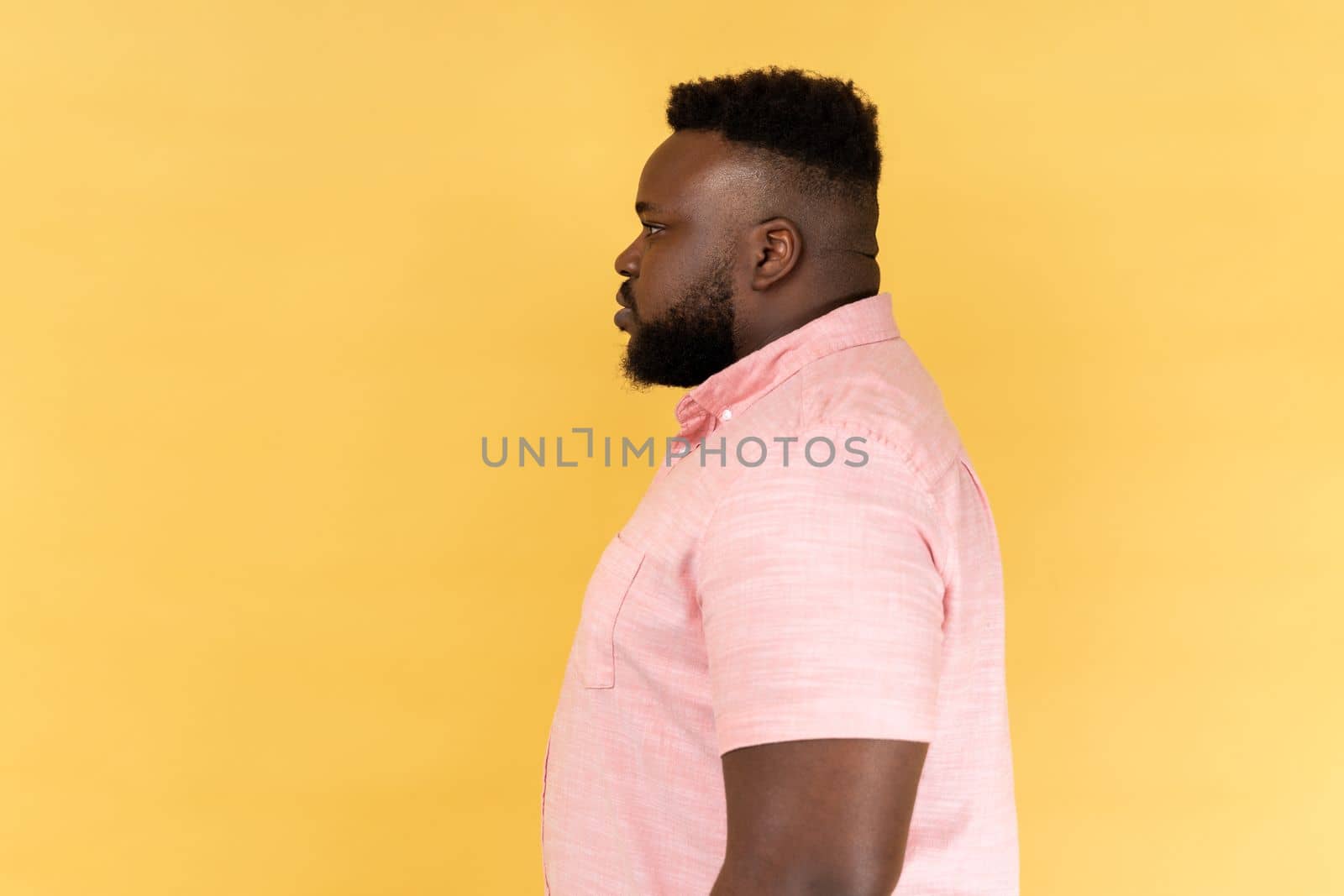 Portrait of serious strict young adult man wearing pink shirt standing and looking ahead with bossy facial expression, being in bad mood. Indoor studio shot isolated on yellow background.