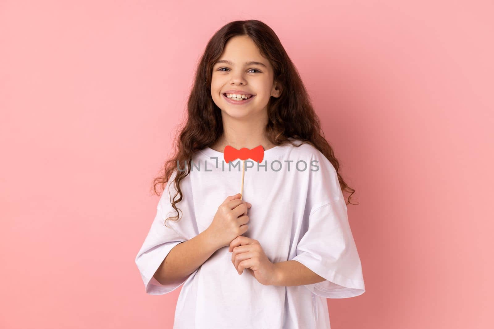 Portrait of satisfied little girl wearing white T-shirt holding funny paper bow tie, having fun playing game, wearing masquerade accessory. Indoor studio shot isolated on pink background.