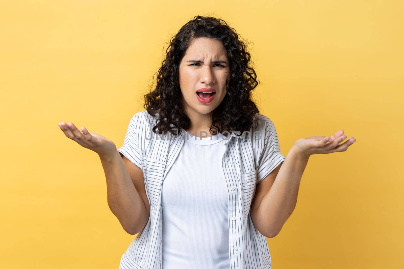 Portrait of unhappy stressed beautiful woman with dark wavy hair standing raised arms and asking, expressing negative emotions. Indoor studio shot isolated on yellow background.
