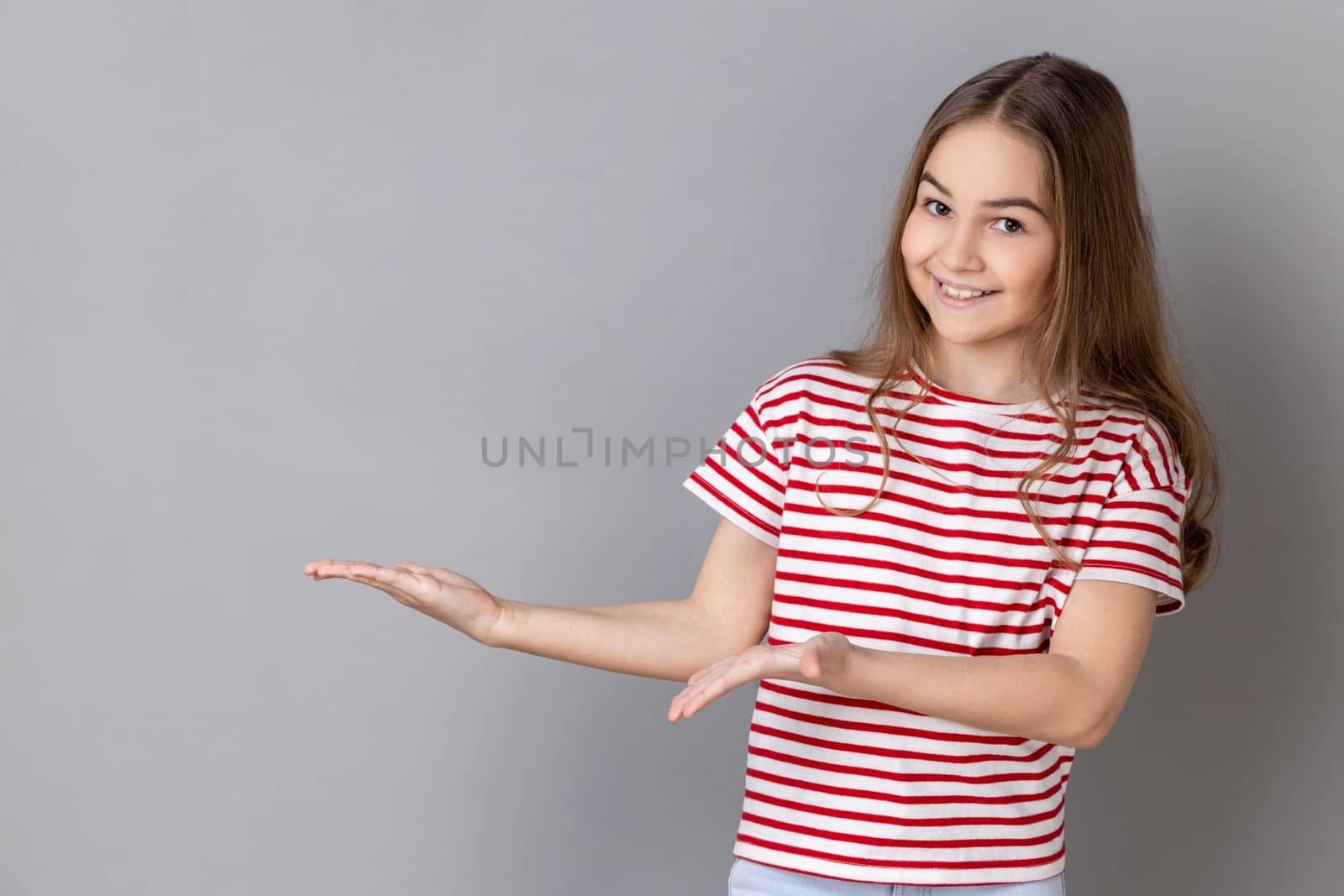 Portrait of generous charming little girl wearing striped T-shirt showing welcome gesture and empty space on wall for your best advertising. Indoor studio shot isolated on gray background.