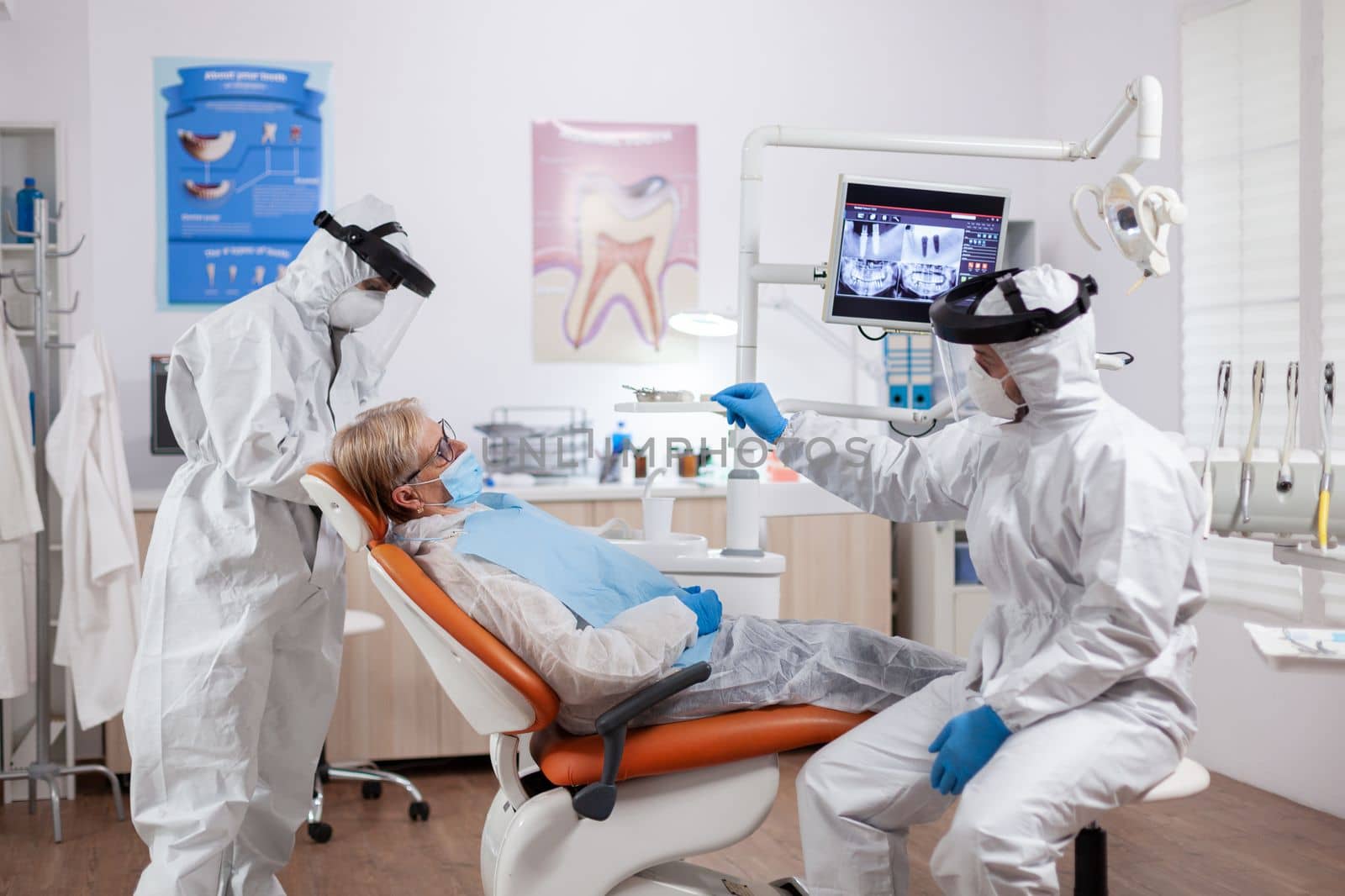 Dentist wearing equipment agasint coronavirus dicussing about teeth hygiene with senior patient. Elderly woman in protective uniform during medical examination in dental clinic.