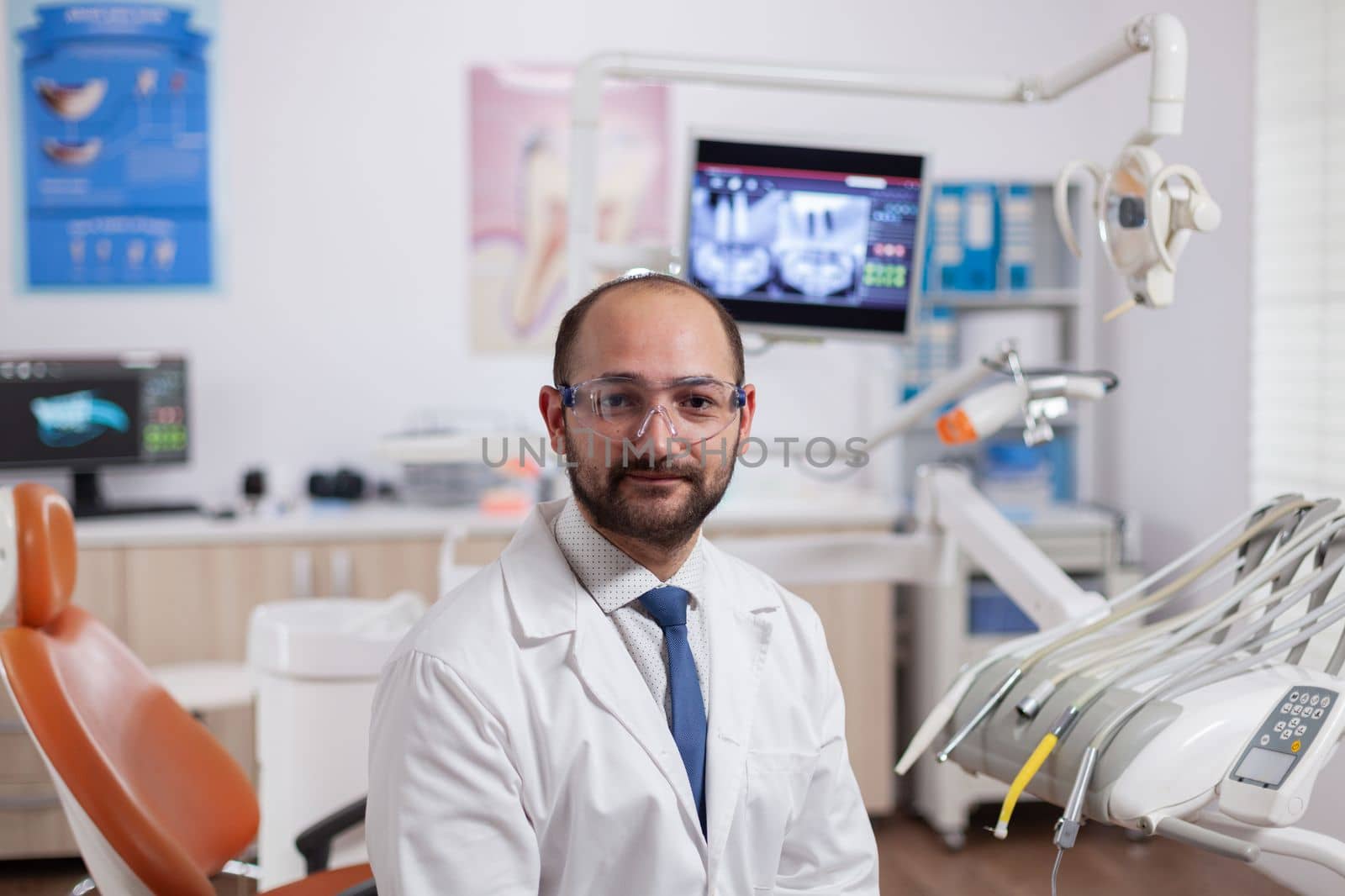 Confident dentist in stomatology cabinet with orange equiptment wearing dental uniform. Medical specialist in oral hygiene wearing lab coat looking at camera in dentistry office.