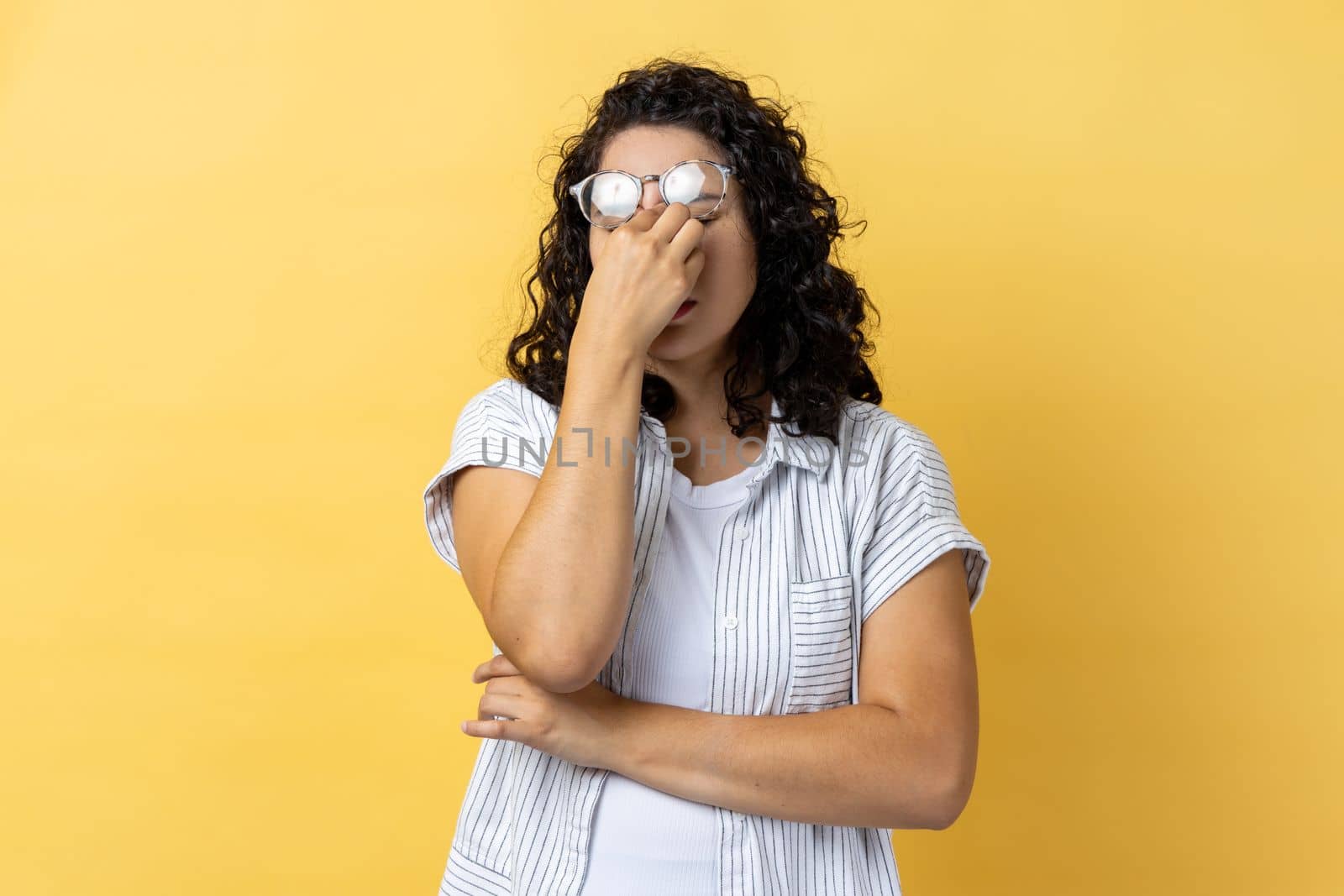 Portrait of tired exhausted attractive young adult woman with dark wavy hair standing with raised eyeglasses, feels pain in eyes. Indoor studio shot isolated on yellow background.
