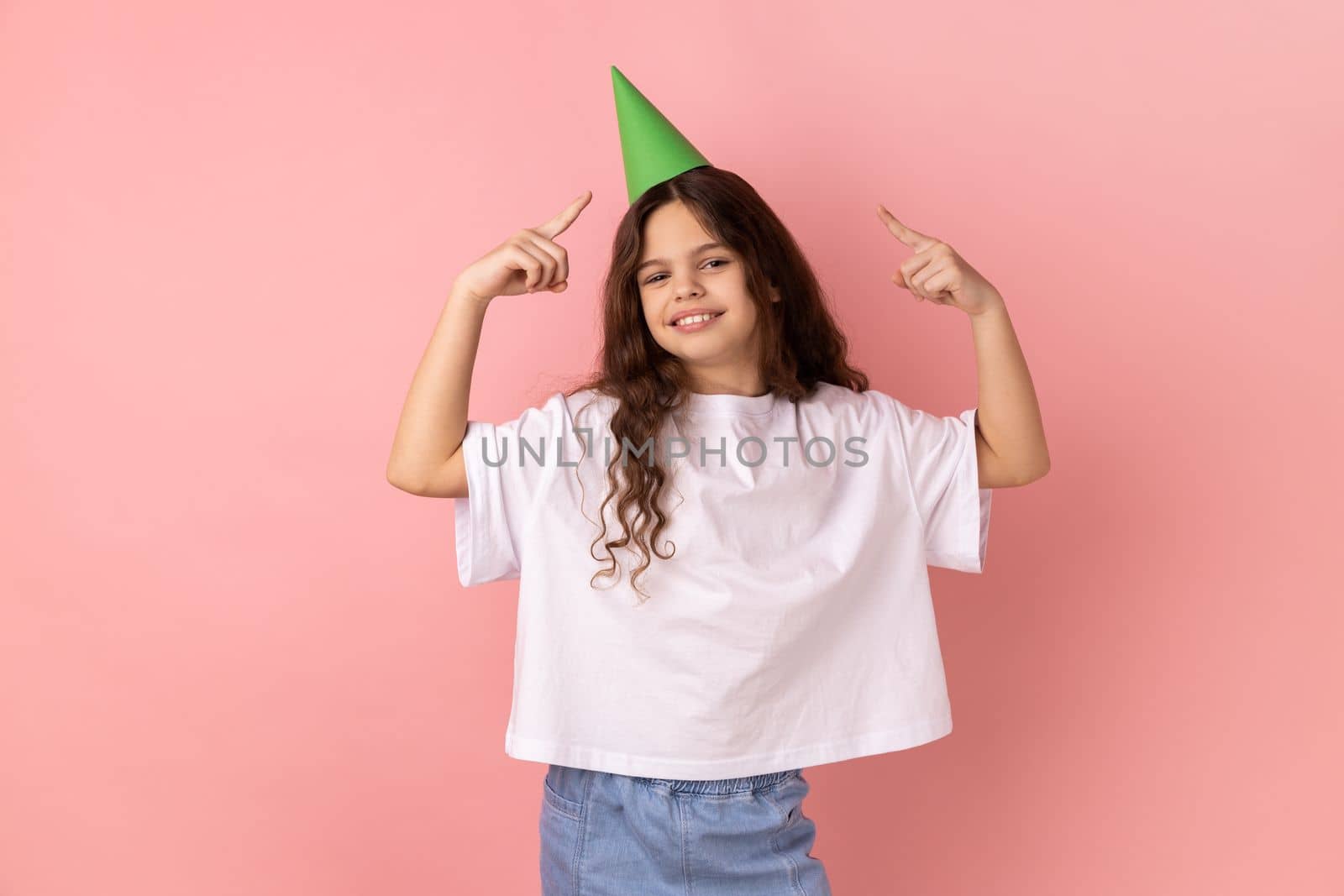 Portrait of little girl wearing white T-shirt pointing at party cone on her head and looking at camera with toothy smile, celebrating birthday. Indoor studio shot isolated on pink background.