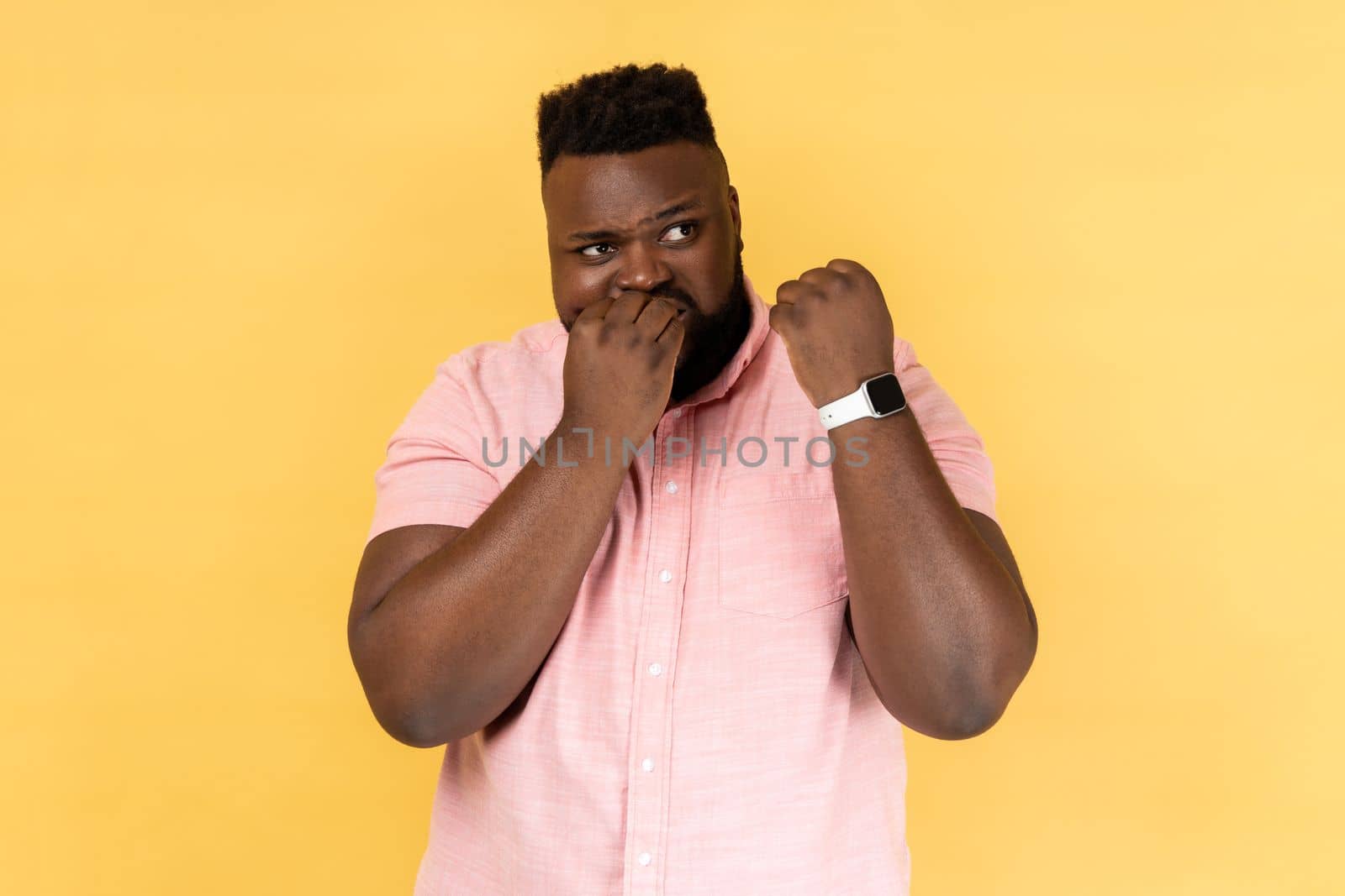 Portrait of nervous bearded man wearing pink shirt showing her smart watch and biting his finger nails, has not enough time. Indoor studio shot isolated on yellow background.