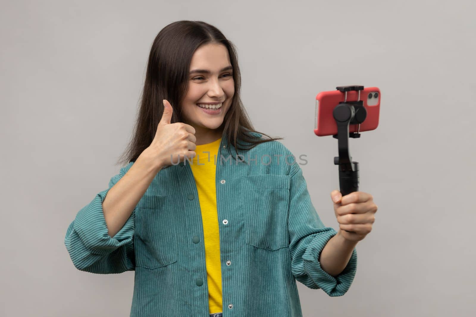Beautiful woman holding smartphone on steadycam in hand, blogger broadcasting livestream or making selfie, showing thumb up, wearing casual style jacket. Indoor studio shot isolated on gray background