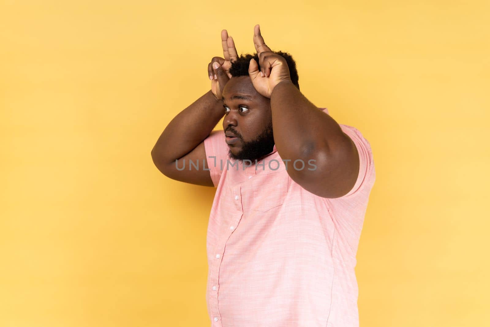 Side view of man wearing pink shirt showing bull horn gesture, looking at camera with hostile angry expression, bully male threatening to attack. Indoor studio shot isolated on yellow background.