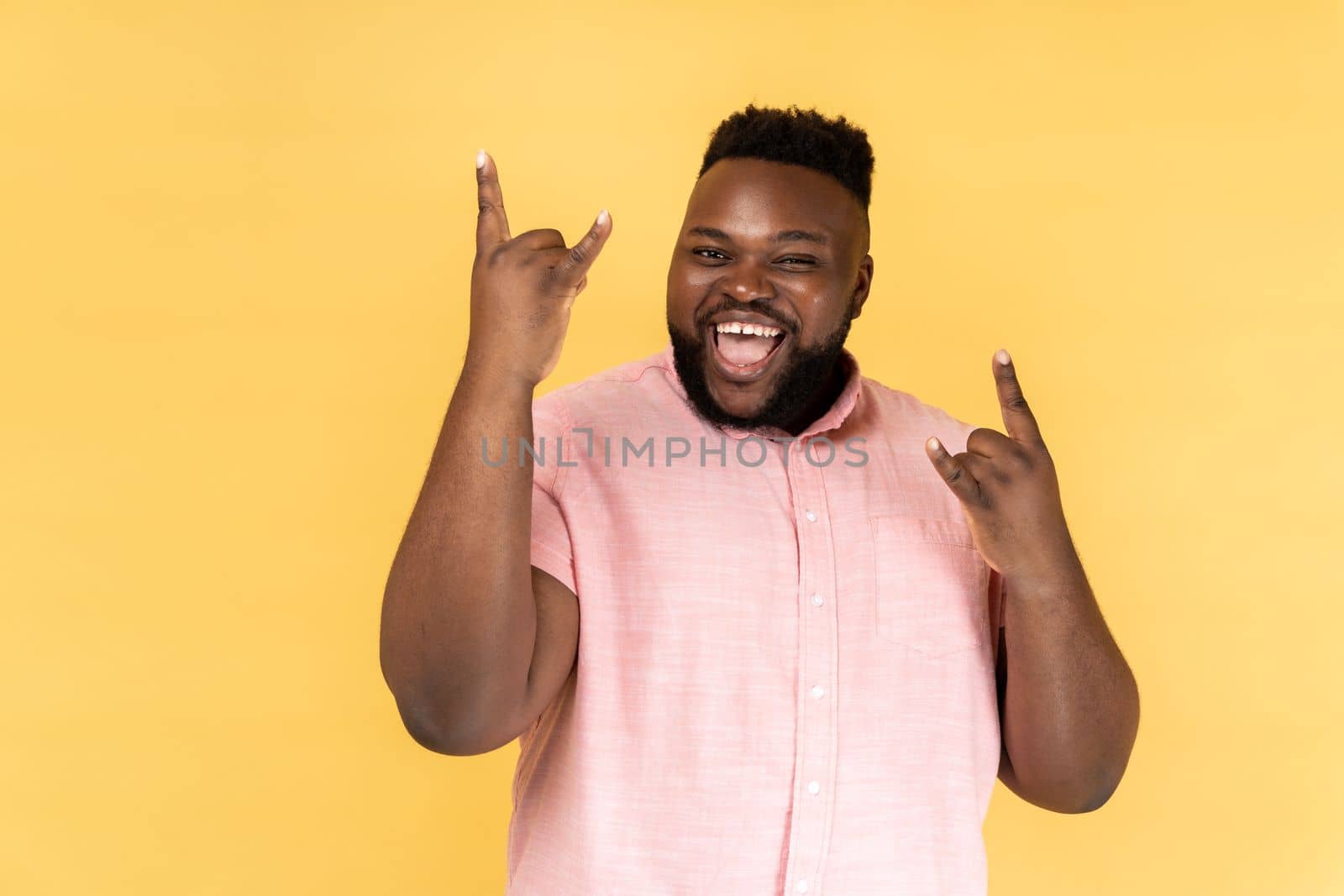 Portrait of disobedient man wearing pink shirt showing rock and roll gesture and excitedly screaming, having fun, enjoying heavy music. Indoor studio shot isolated on yellow background.