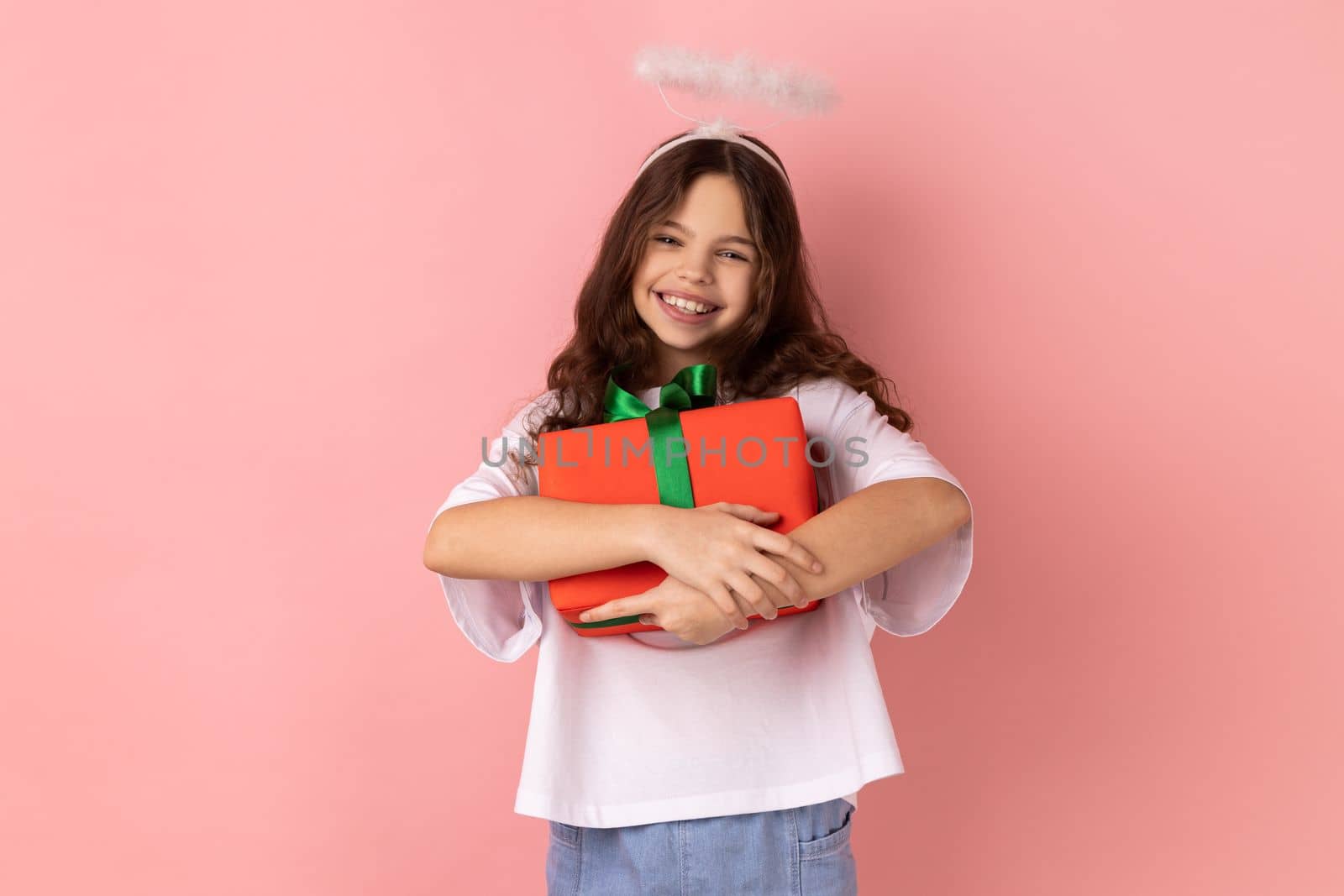 Portrait of little girl wearing white T-shirt and angelic halo embracing wrapped box, enjoying birthday gift, satisfied with long-awaited present. Indoor studio shot isolated on pink background.