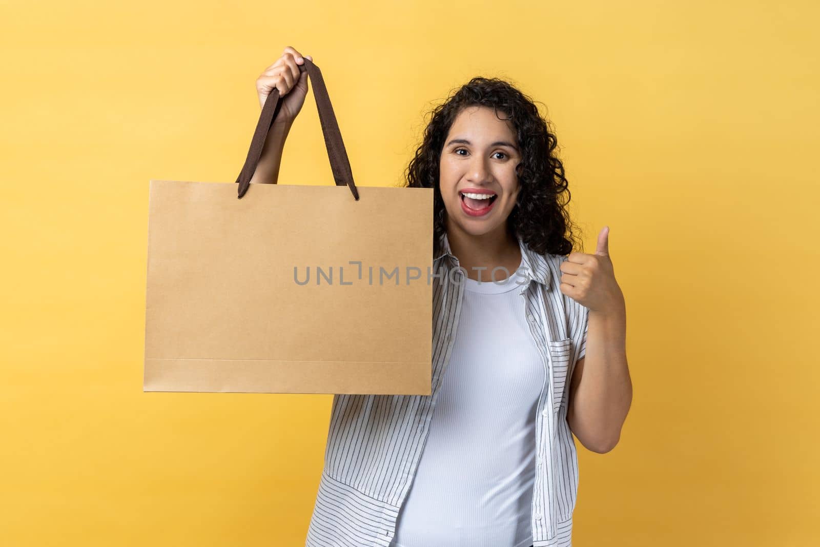 Portrait of satisfied positive woman with dark wavy hair standing with shopping bag and showing thumb up, recommend mall for shopping. Indoor studio shot isolated on yellow background.