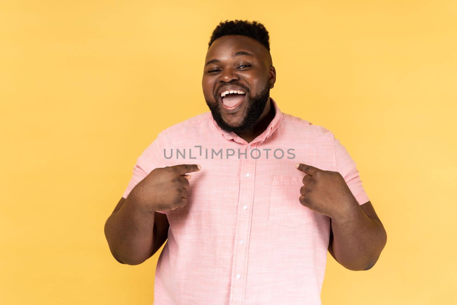 This is me. Portrait of man wearing pink shirt pointing finger at himself feeling proud and ambitious, boasting successful achievement. Indoor studio shot isolated on yellow background.