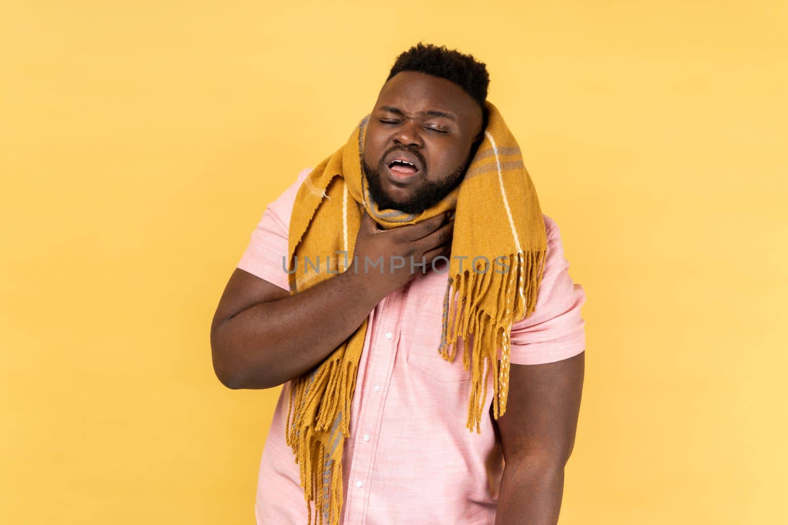 Portrait of man wearing pink shirt in warm scarf feeling unwell with sore throat, high temperature fever, seasonal influenza symptoms. Indoor studio shot isolated on yellow background.