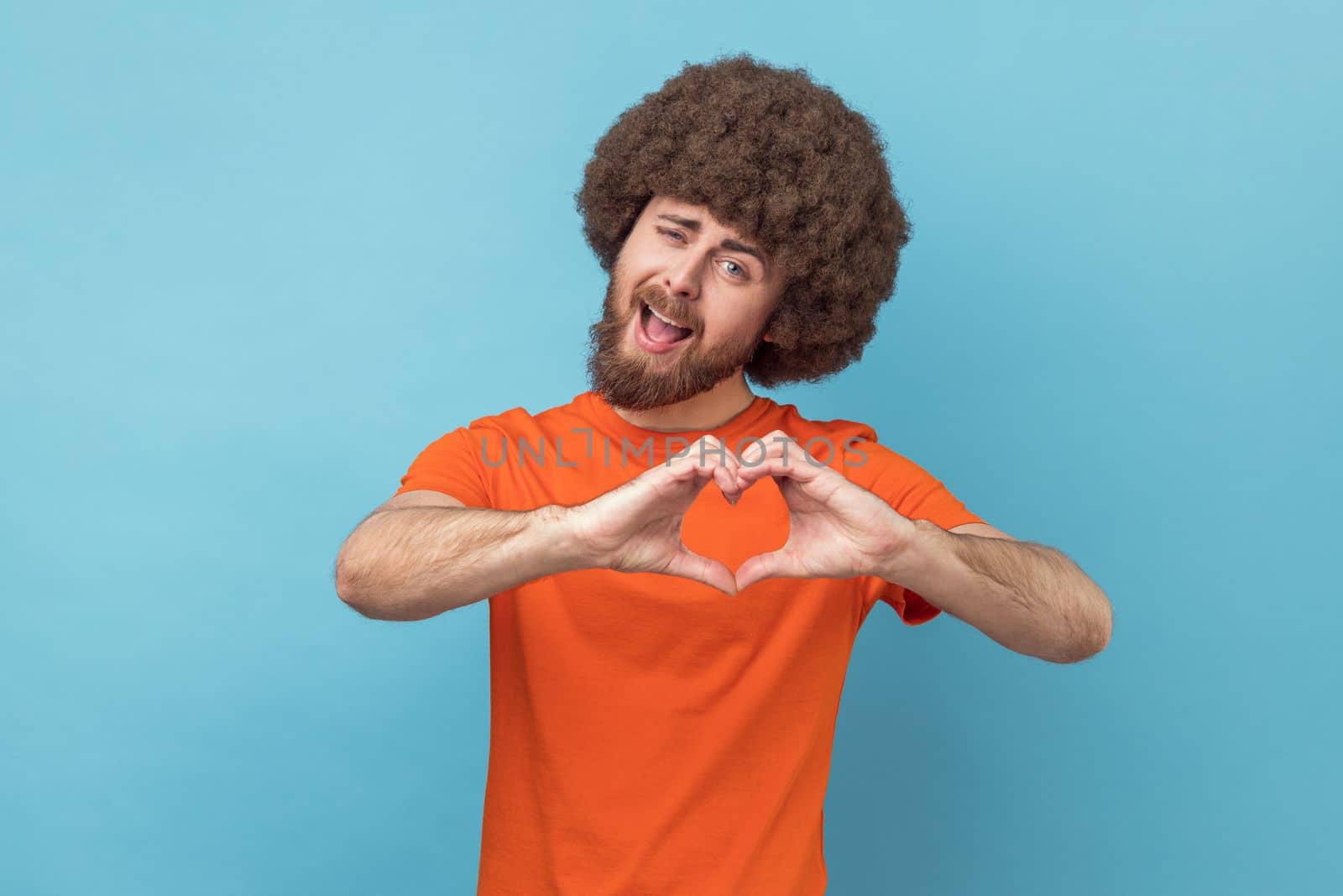 Portrait of romantic man with Afro hairstyle wearing orange T-shirt standing with heart or love gesture and looking at camera with toothy smile. Indoor studio shot isolated on blue background.