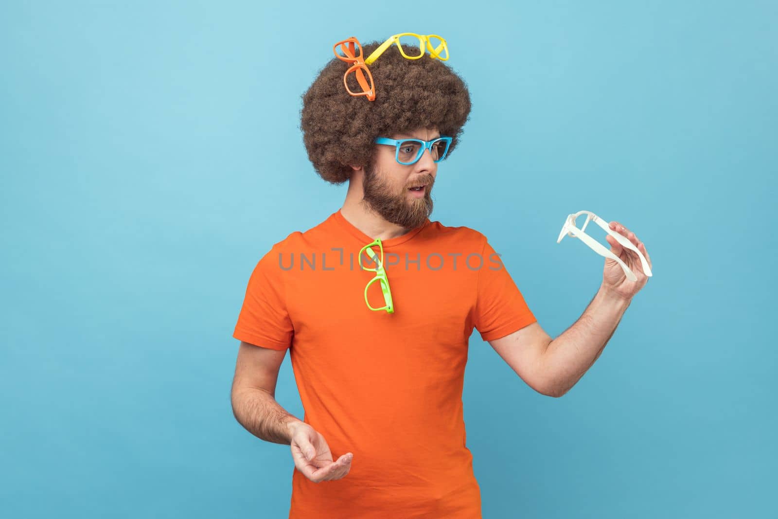 Portrait of funny man with Afro hairstyle wearing orange T-shirt choosing colorful eyeglasses, holding white spectacles, choosing the best color. Indoor studio shot isolated on blue background.