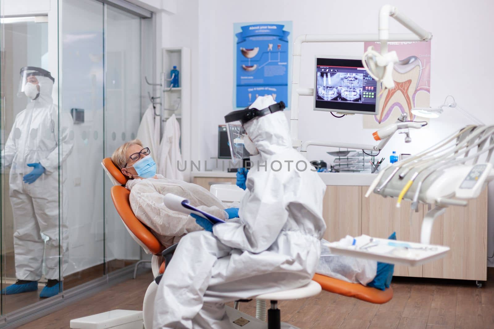 Dentist assitatnt questioning patient wearing protective equipment against coronavirus. Elderly woman in protective uniform during medical examination in dental clinic.