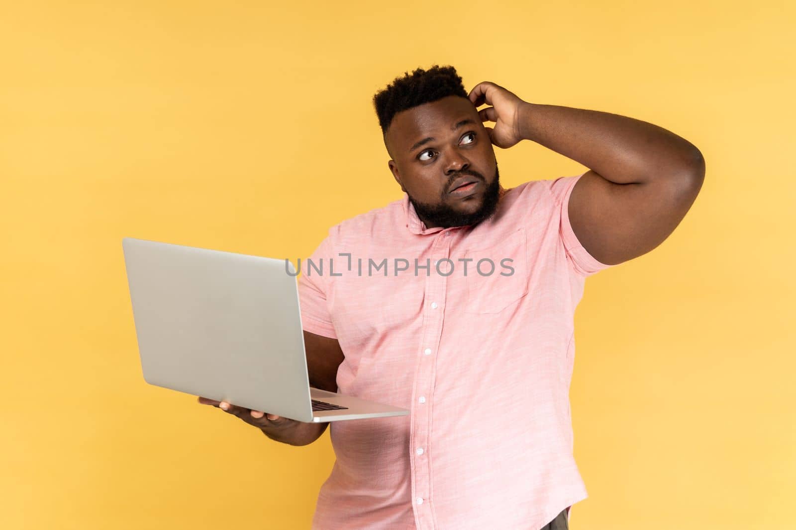 Portrait of thoughtful bearded man wearing pink shirt holding laptop and thinking over startup strategy with serious doubtful expression. Indoor studio shot isolated on yellow background.