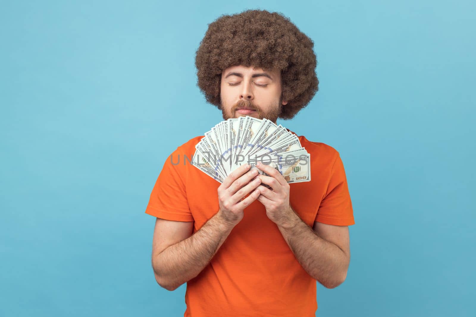 Portrait of man with Afro hairstyle wearing orange T-shirt smelling dollar banknotes with pleasure expression, enjoying big money profit and rich life. Indoor studio shot isolated on blue background.