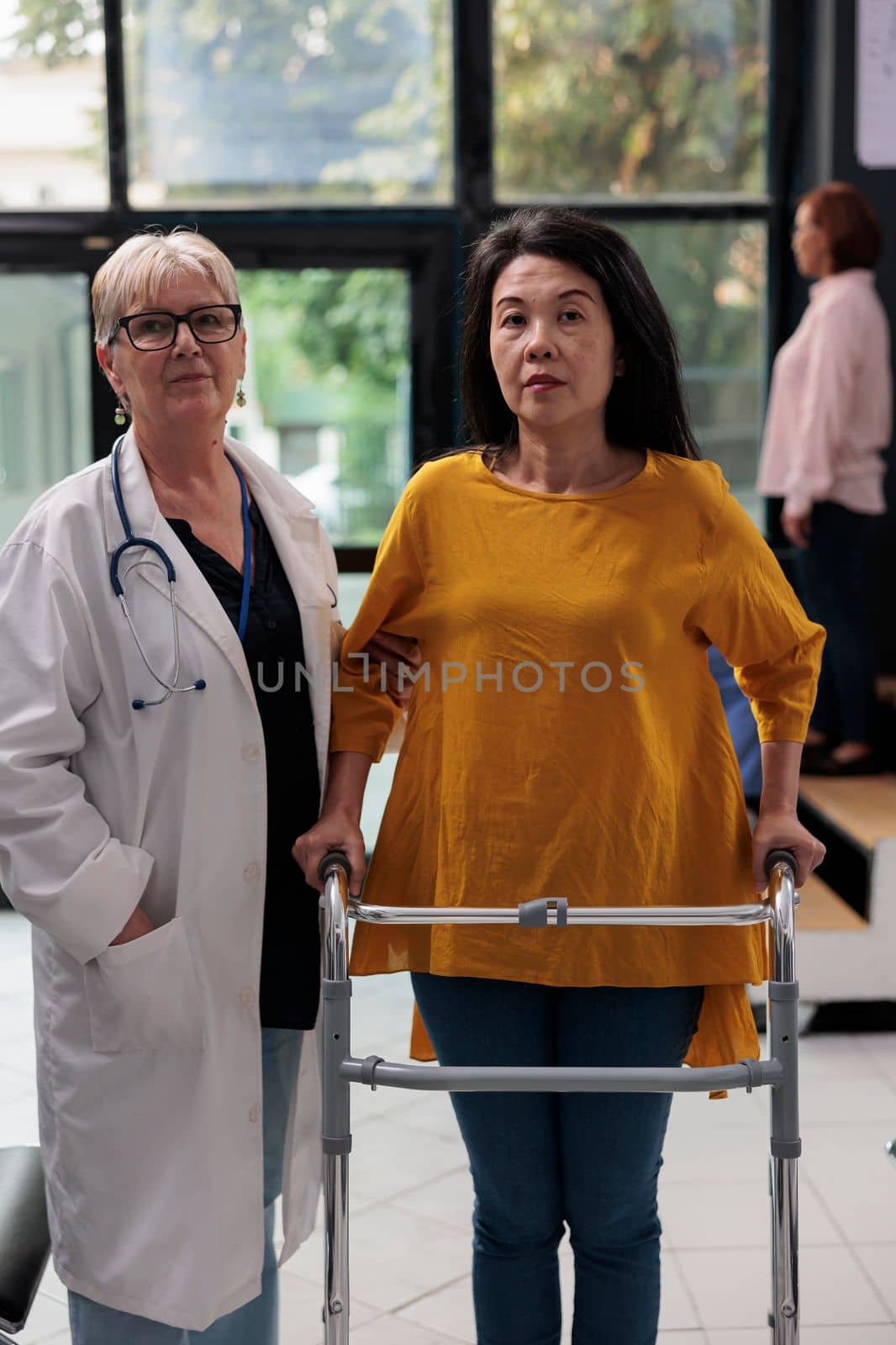 Portrait of medic doing physical therapy exam with asian patient, supporting with walking frame to recover after leg fracture injury. Injured woman attending medical physiotherapy consultation.