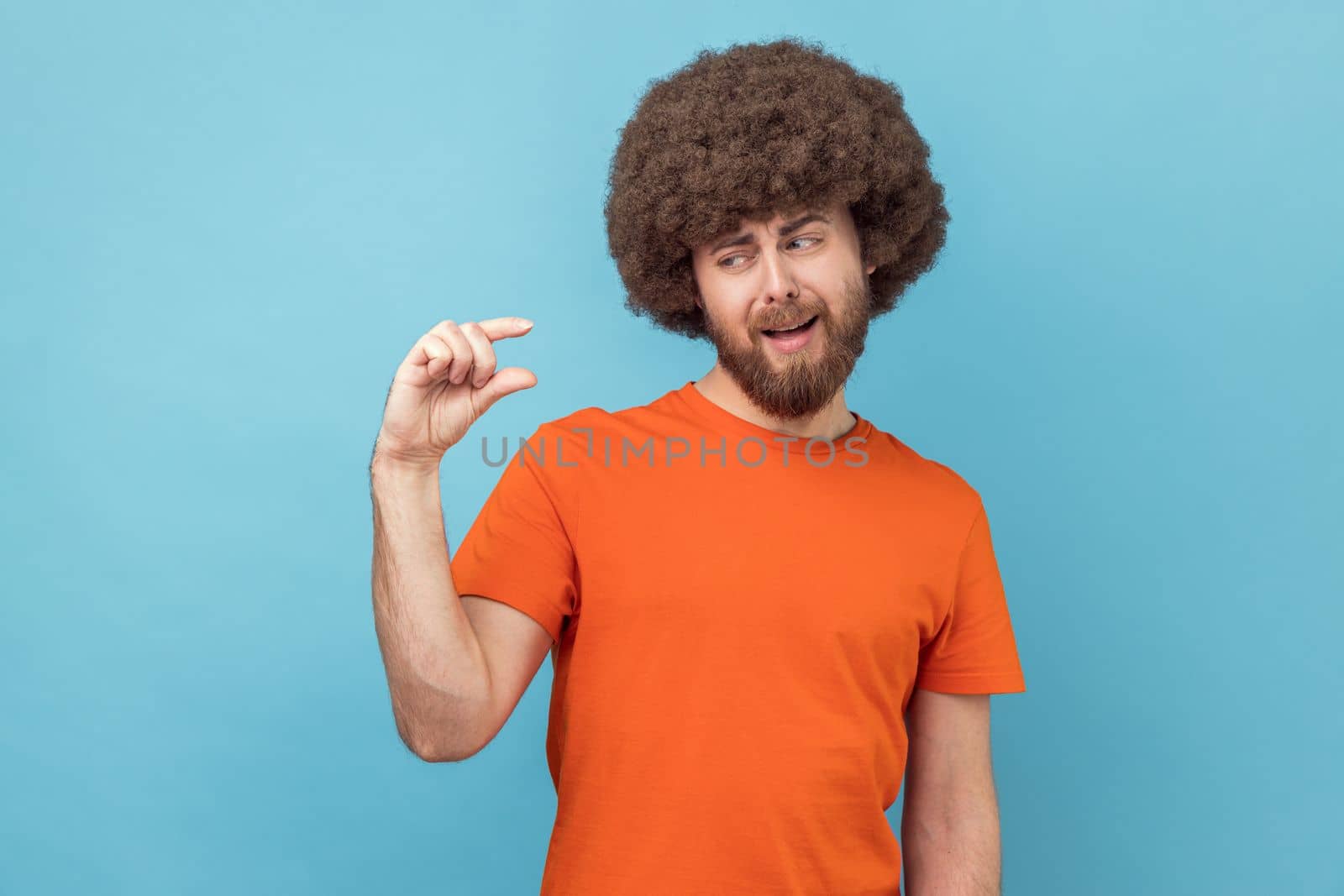 Not very much. Portrait of man with Afro hairstyle wearing orange T-shirt shapes little gesture, demonstrates something very tiny, measures small size. Indoor studio shot isolated on blue background.