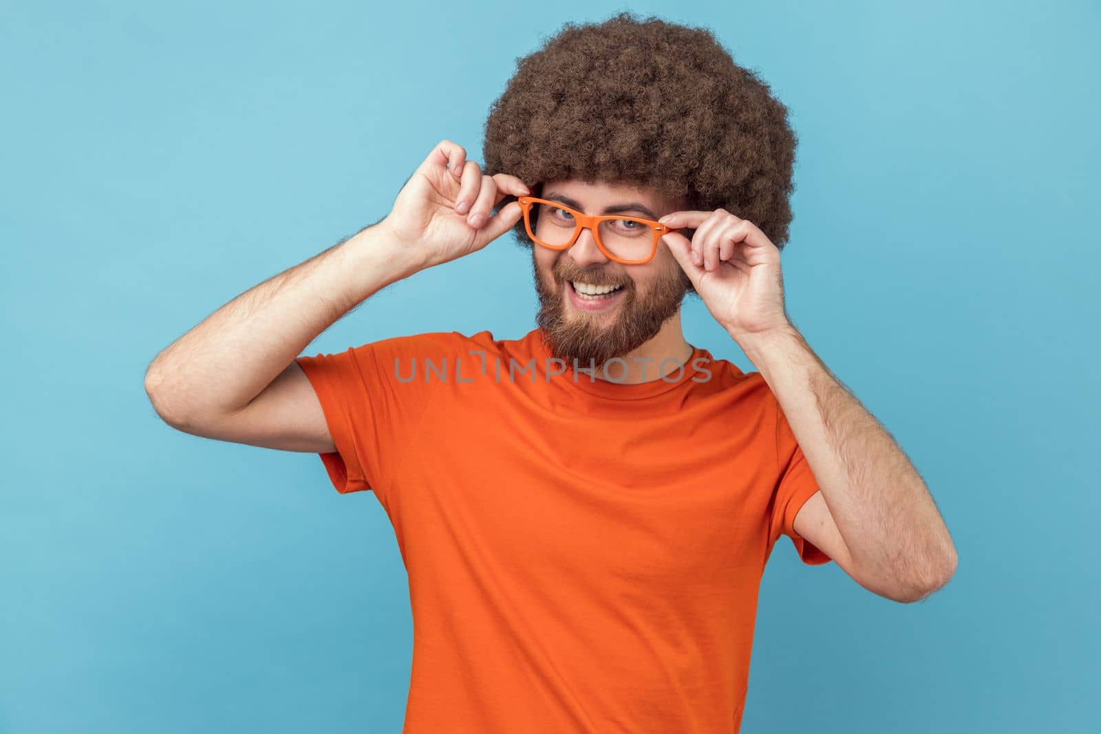 Delighted satisfied man with Afro hairstyle in orange T-shirt admires eyeglasses, standing in a new spectacles, looking at camera with toothy smile. Indoor studio shot isolated on blue background.